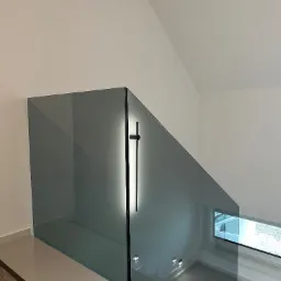 Szklana balustrada schodowa z oświetleniem LED w Wieliczce. Minimalistyczny design, widok z góry na schody z szarymi stopniami i okno z widokiem na zewnątrz.