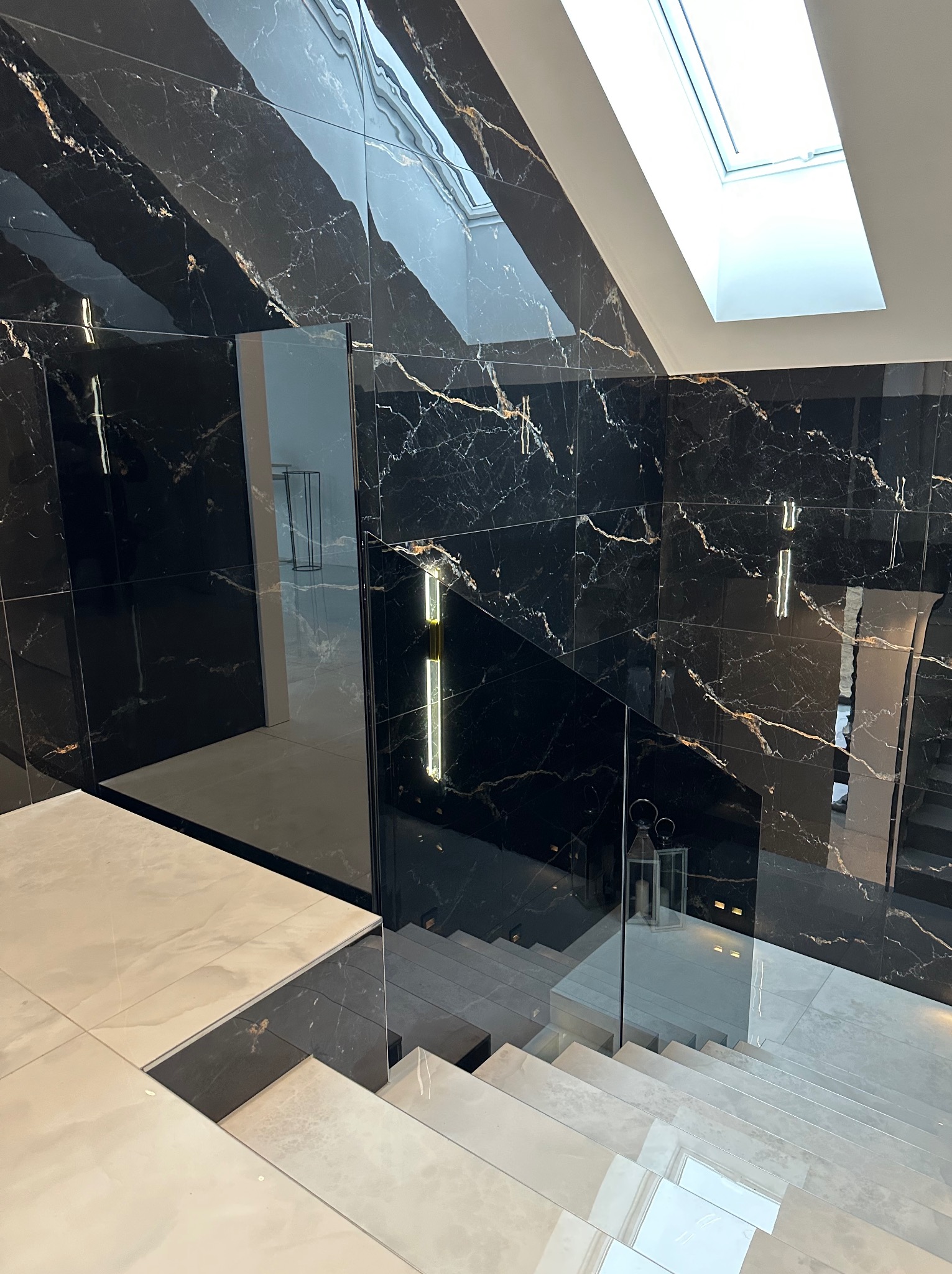 Ekskluzywne schody wewnętrzne z jasnymi stopniami i szklaną balustradą na tle czarnej ściany z marmurowym wzorem, oświetlone światłem z okna dachowego.