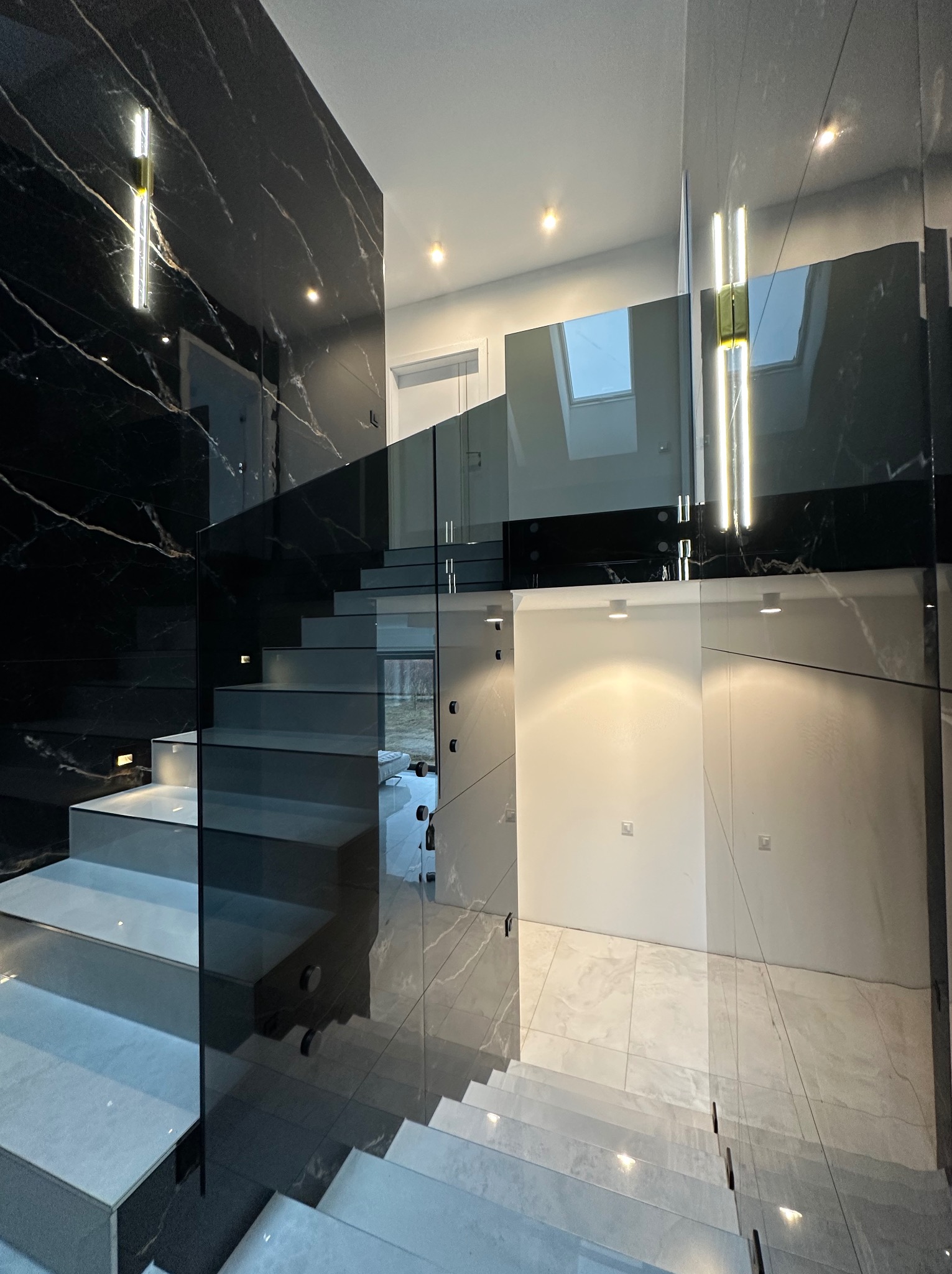 Nowoczesne schody z szarymi stopniami i szklaną balustradą, oświetlone punktowymi lampami, z czarną marmurową ścianą w tle.