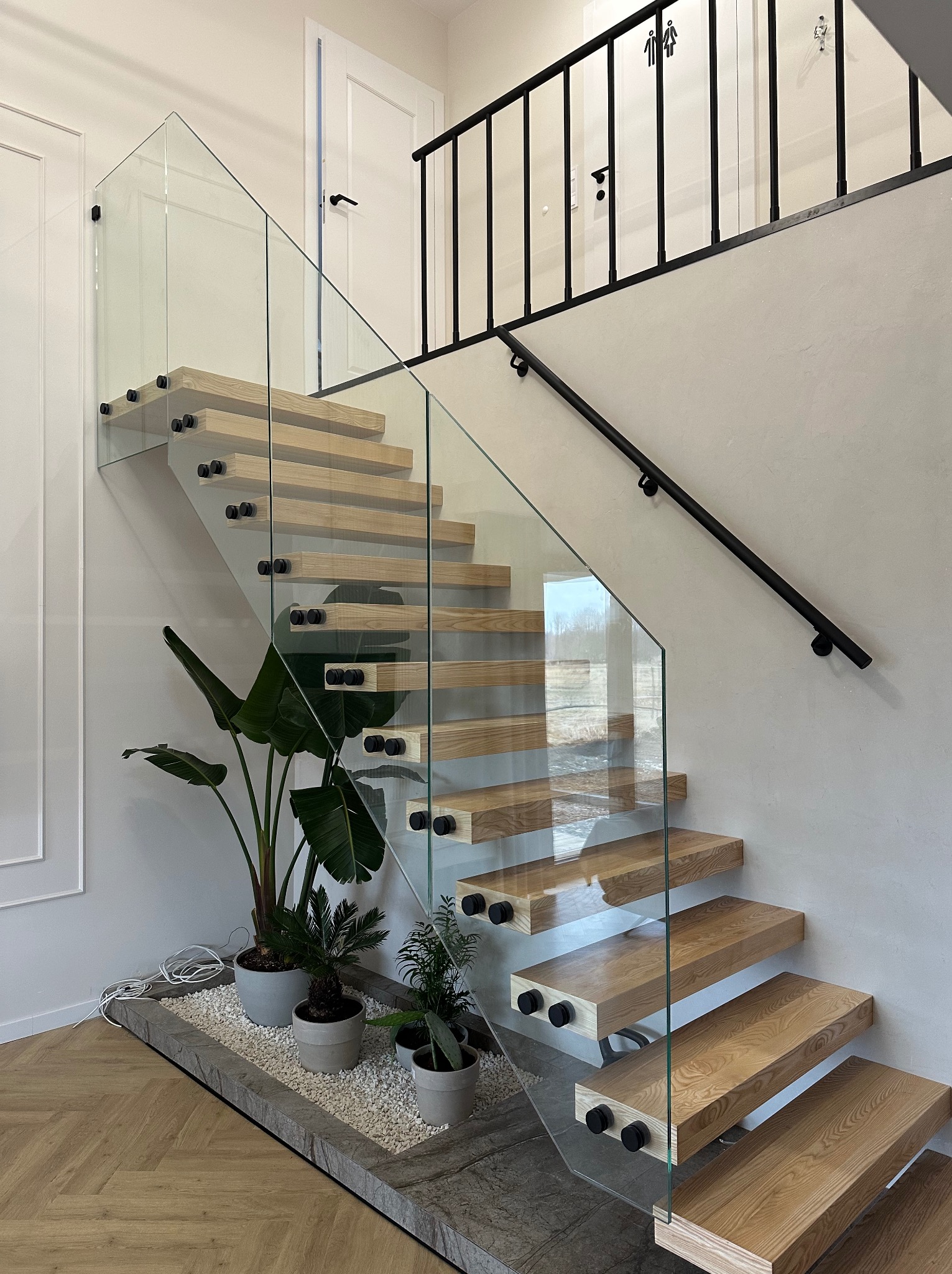 Nowoczesne schody drewniane z przezroczystą balustradą szklaną, mocowaną czarnymi uchwytami, z roślinami doniczkowymi u podstawy i czarną poręczą na piętrze.