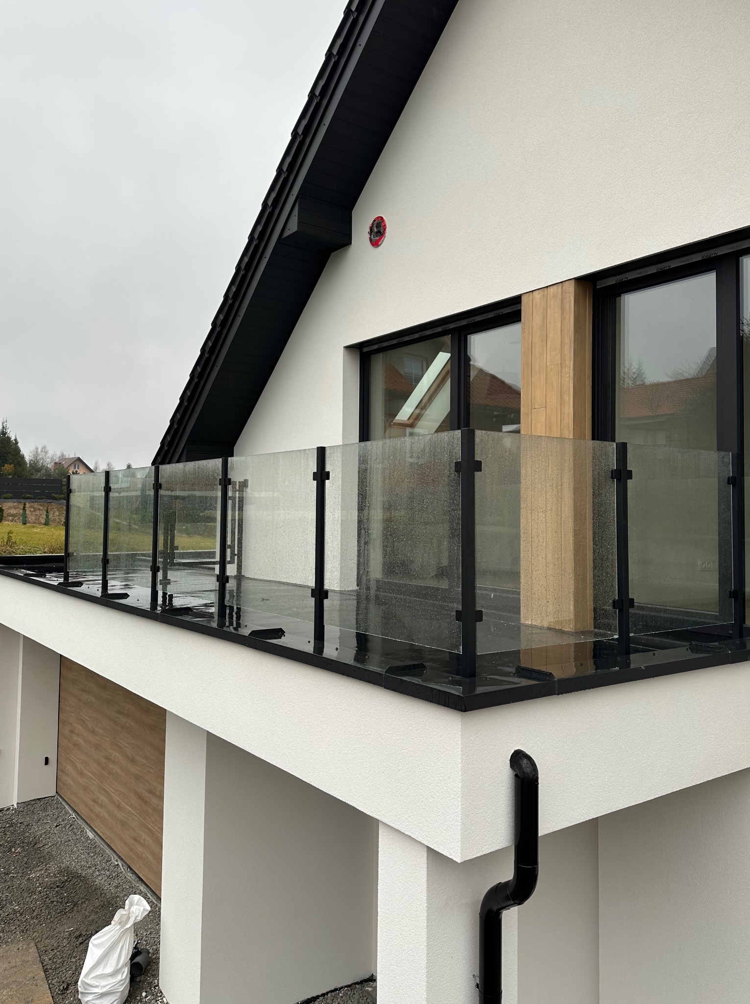 Nowoczesny balkon z czarną ramą i szklanym wypełnieniem, widok z zewnątrz na minimalistyczny dom w pochmurny dzień.