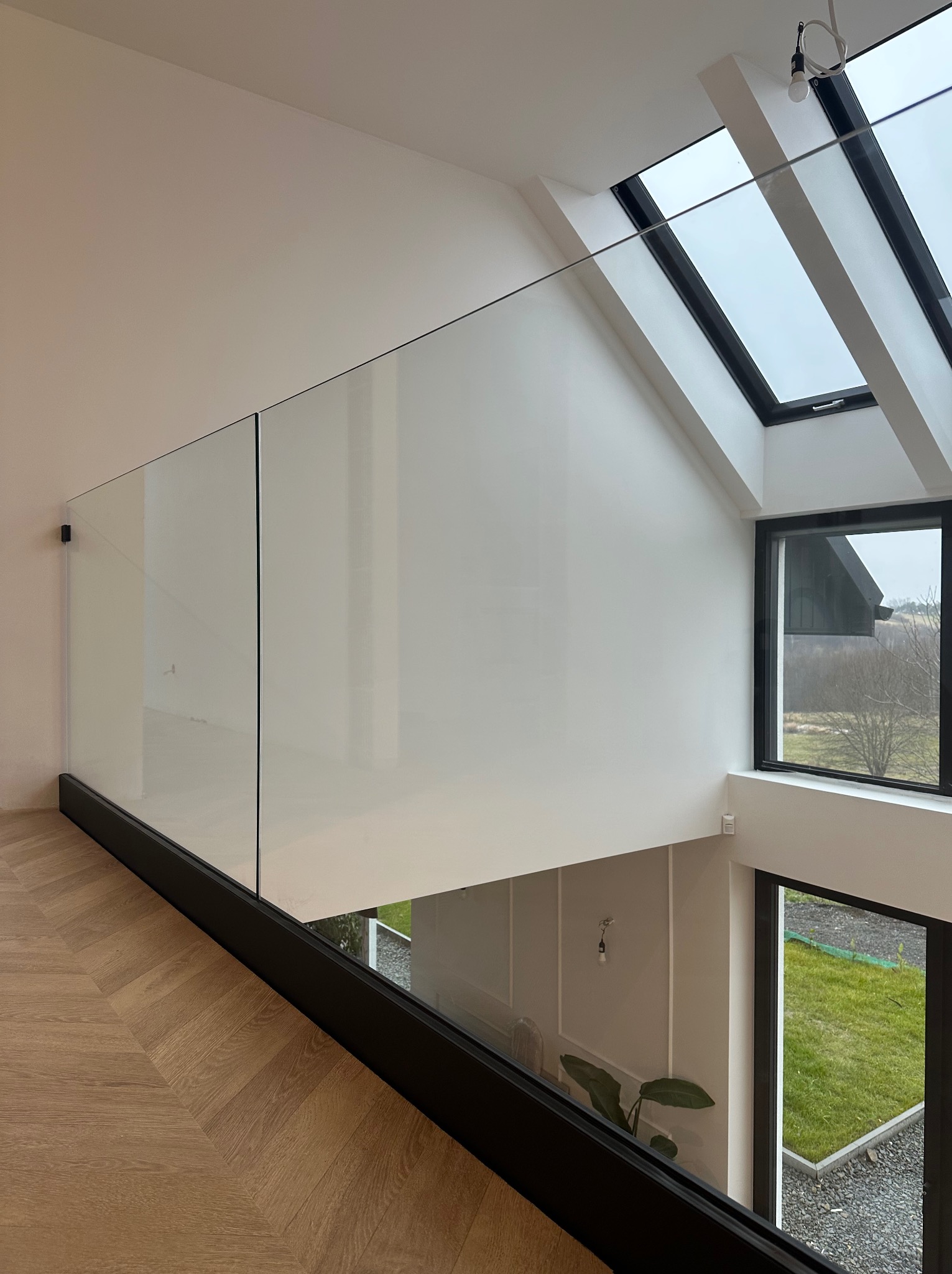 Minimalistyczne wnętrze z przeszkloną balustradą na antresoli, parkiet w jodełkę, widok na ogród przez okno, doświetlone oknami dachowymi.