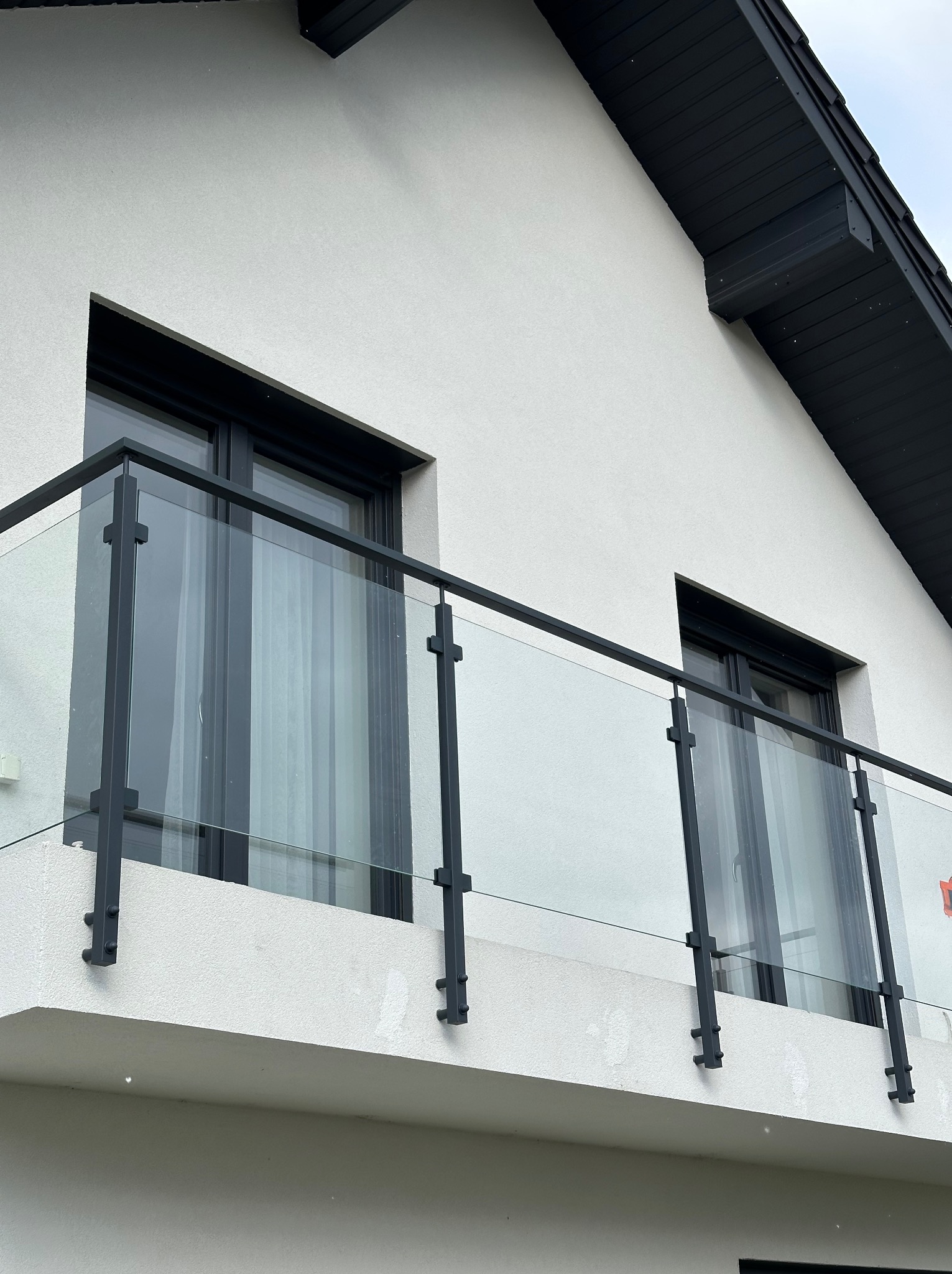 Balkon z nowoczesną szklaną balustradą z czarnymi metalowymi słupkami i ramą, widok z dołu na białą elewację domu z czarnym dachem.