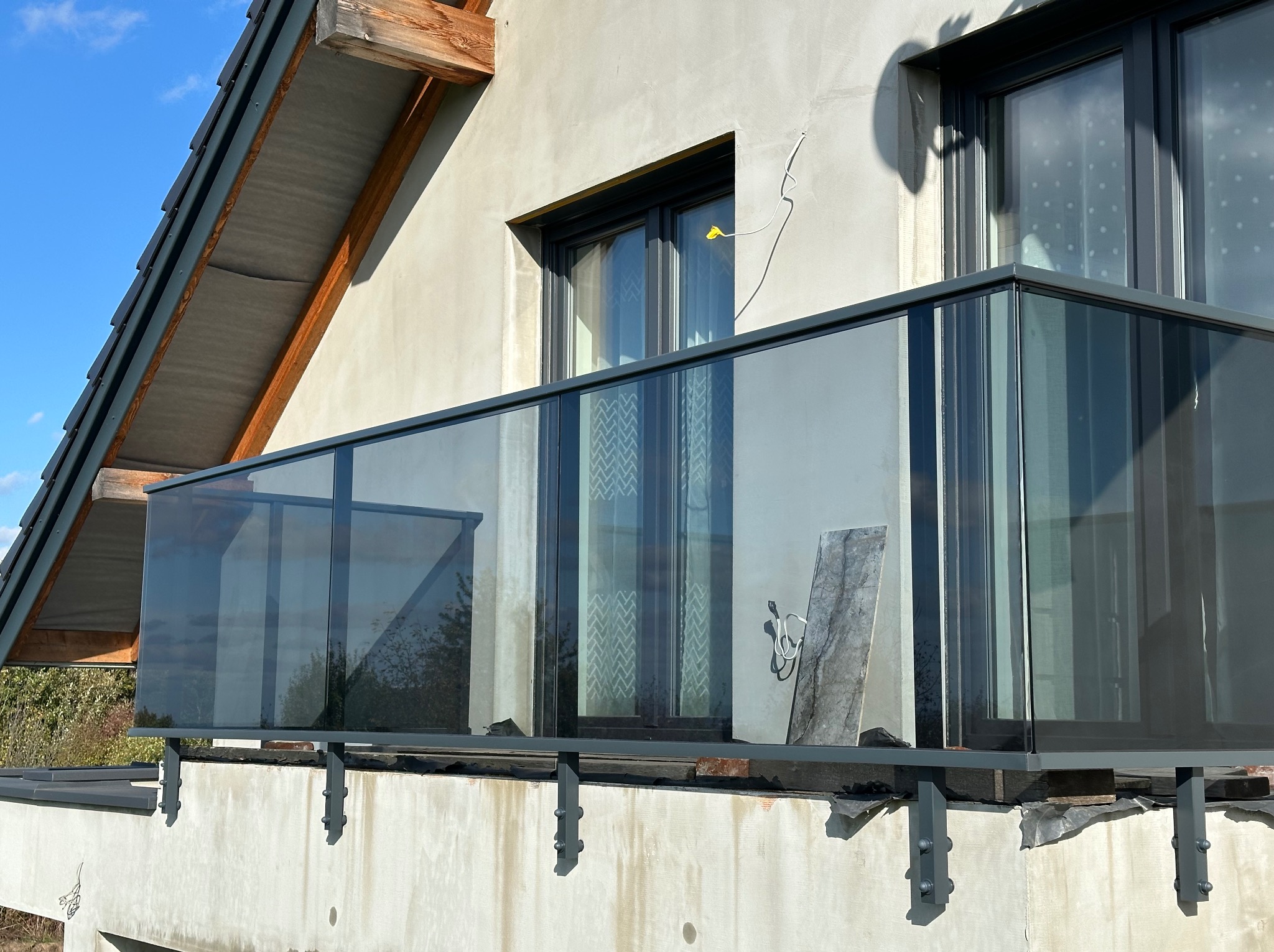 Szklana balustrada balkonowa z ciemną ramą na tle surowej elewacji budynku w trakcie wykończenia.