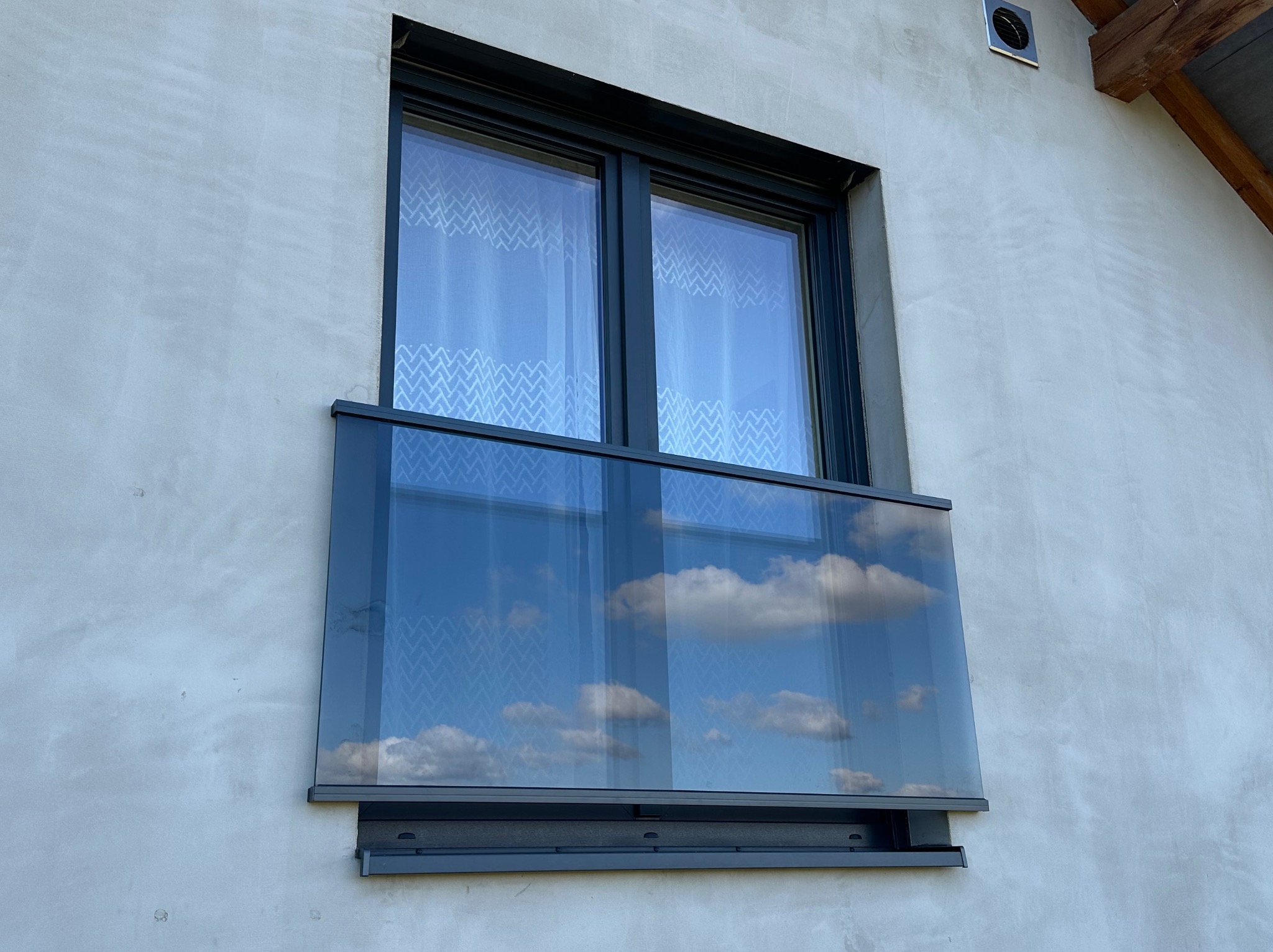 Zewnętrzna balustrada szklana zamontowana przy oknie na elewacji budynku, odbijająca błękitne niebo z chmurami.