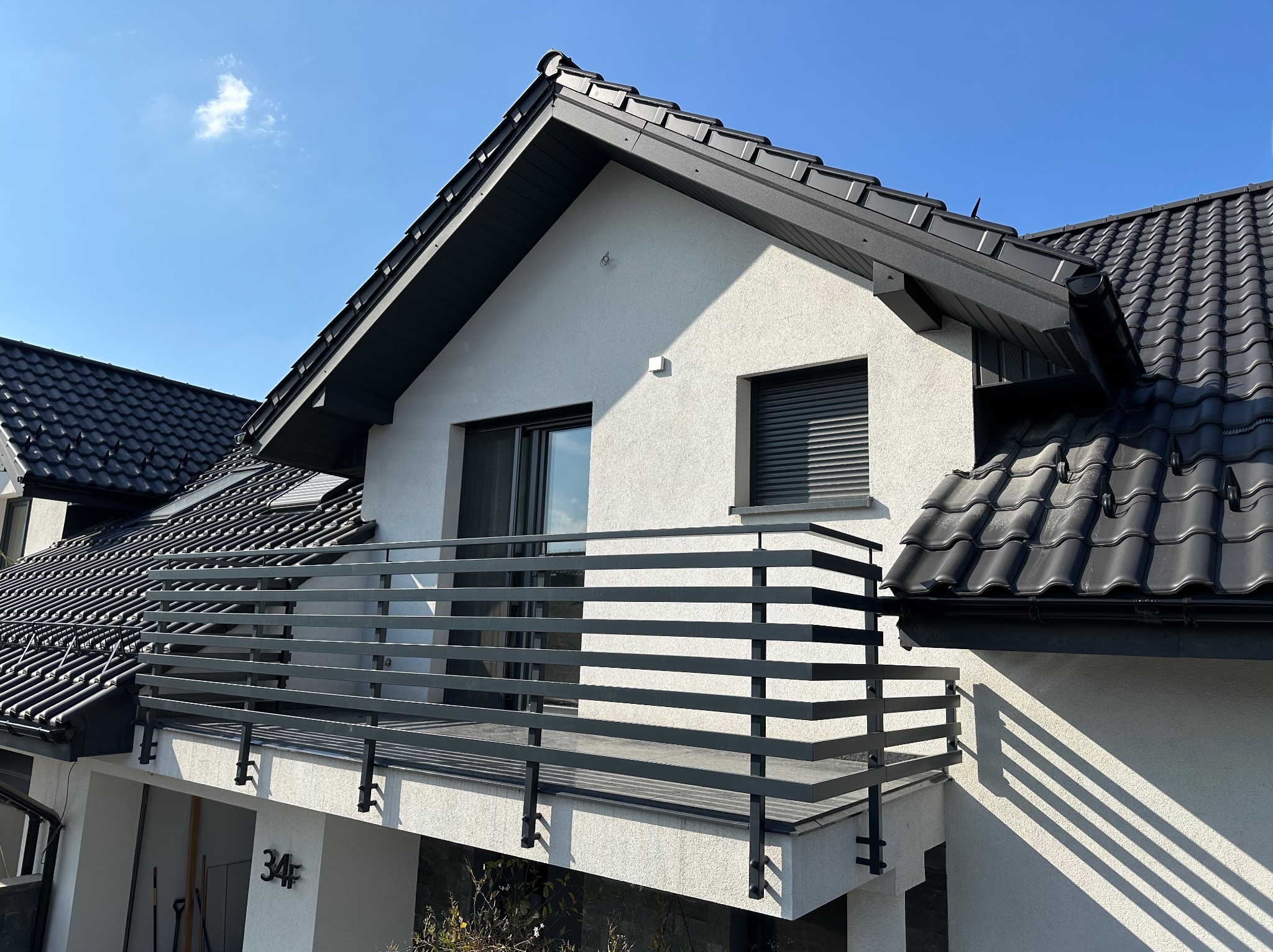 Nowoczesny balkon z czarną, poziomą balustradą na tle białej elewacji domu z czarnym dachem i błękitnym niebem. Ujęcie z dołu, widoczny numer domu.