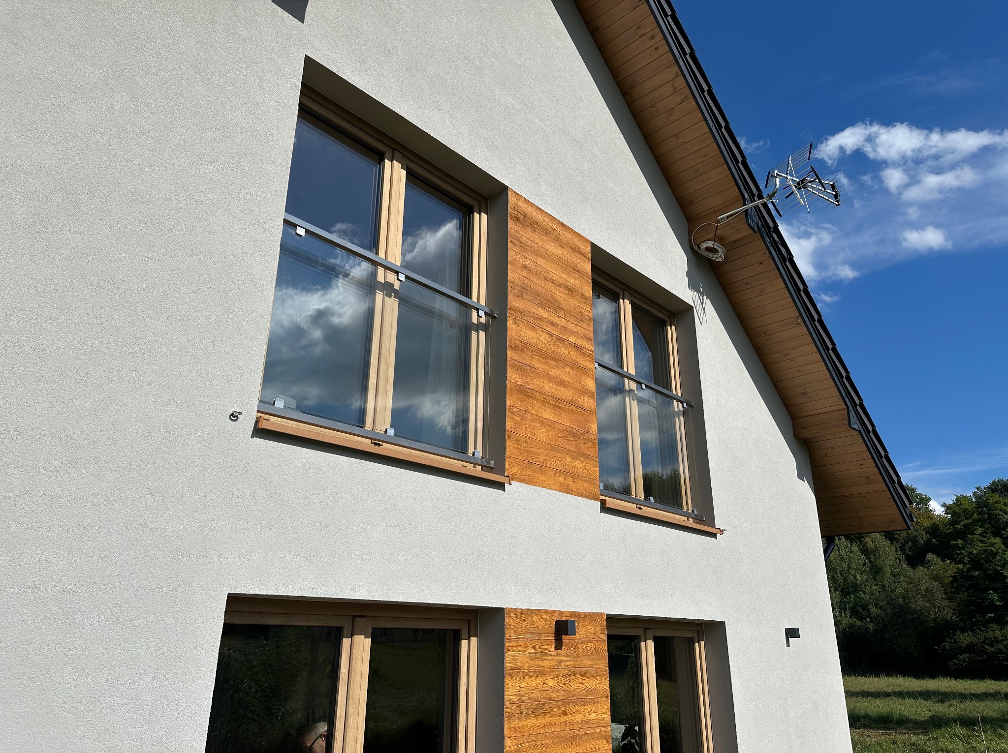 Szklane balustrady balkonowe z drewnianymi elementami na tle nowoczesnego domu z białą elewacją i błękitnym niebem. Detal architektoniczny.