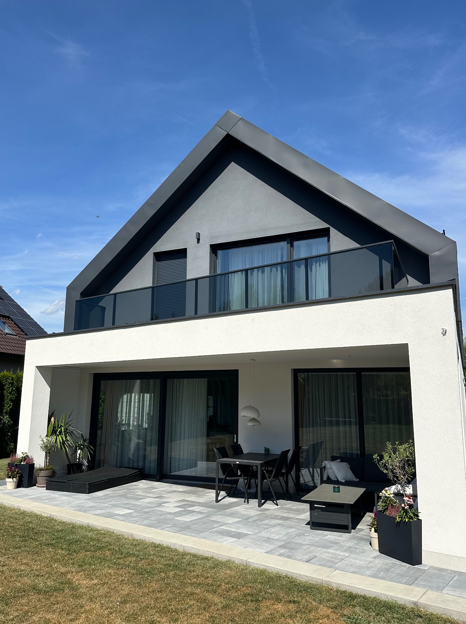 Nowoczesny dom z balkonem zabezpieczonym szklaną balustradą. Minimalistyczny design, taras z meblami ogrodowymi i starannie dobraną roślinnością w donicach.