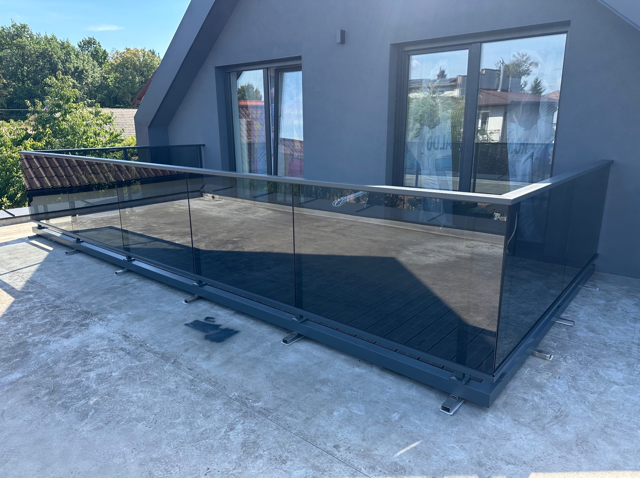 Nowoczesna balustrada ze szkła i szarego metalu na balkonie. Widoczne mocowania. Tło stanowią okna i fragment zieleni, tworząc minimalistyczny, a zarazem elegancki design.