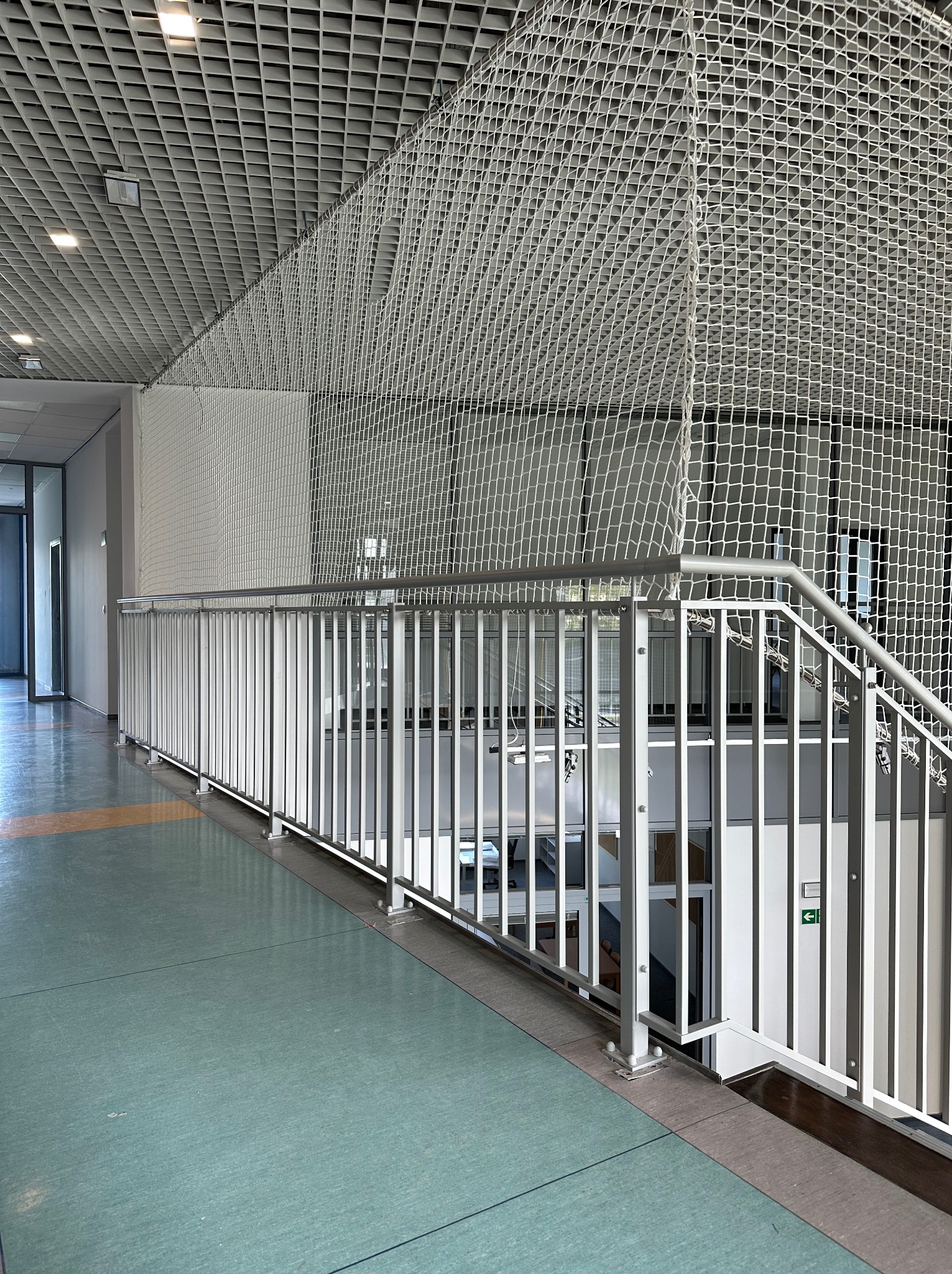 Biała balustrada schodowa z siatką zabezpieczającą w budynku użyteczności publicznej, widok z boku, oświetlenie sufitowe.