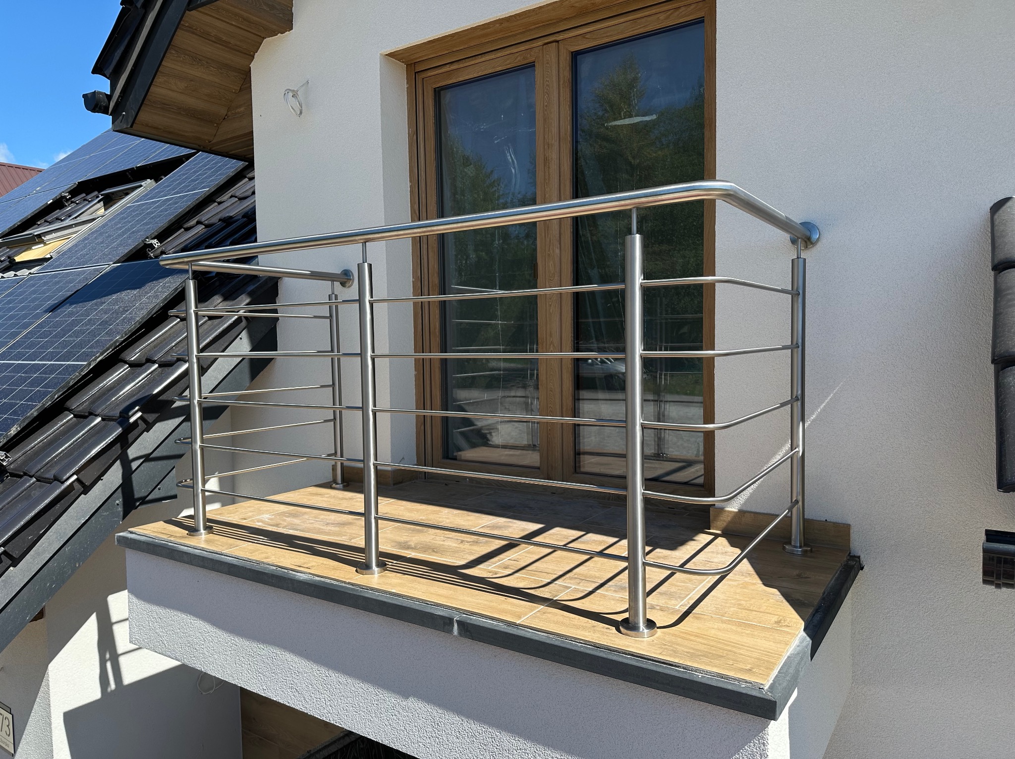 Balkon z balustradą ze stali nierdzewnej, drewnopodobne płytki na podłodze, okno z drewnianą ramą, panele słoneczne na dachu. Nowoczesne wykończenie.