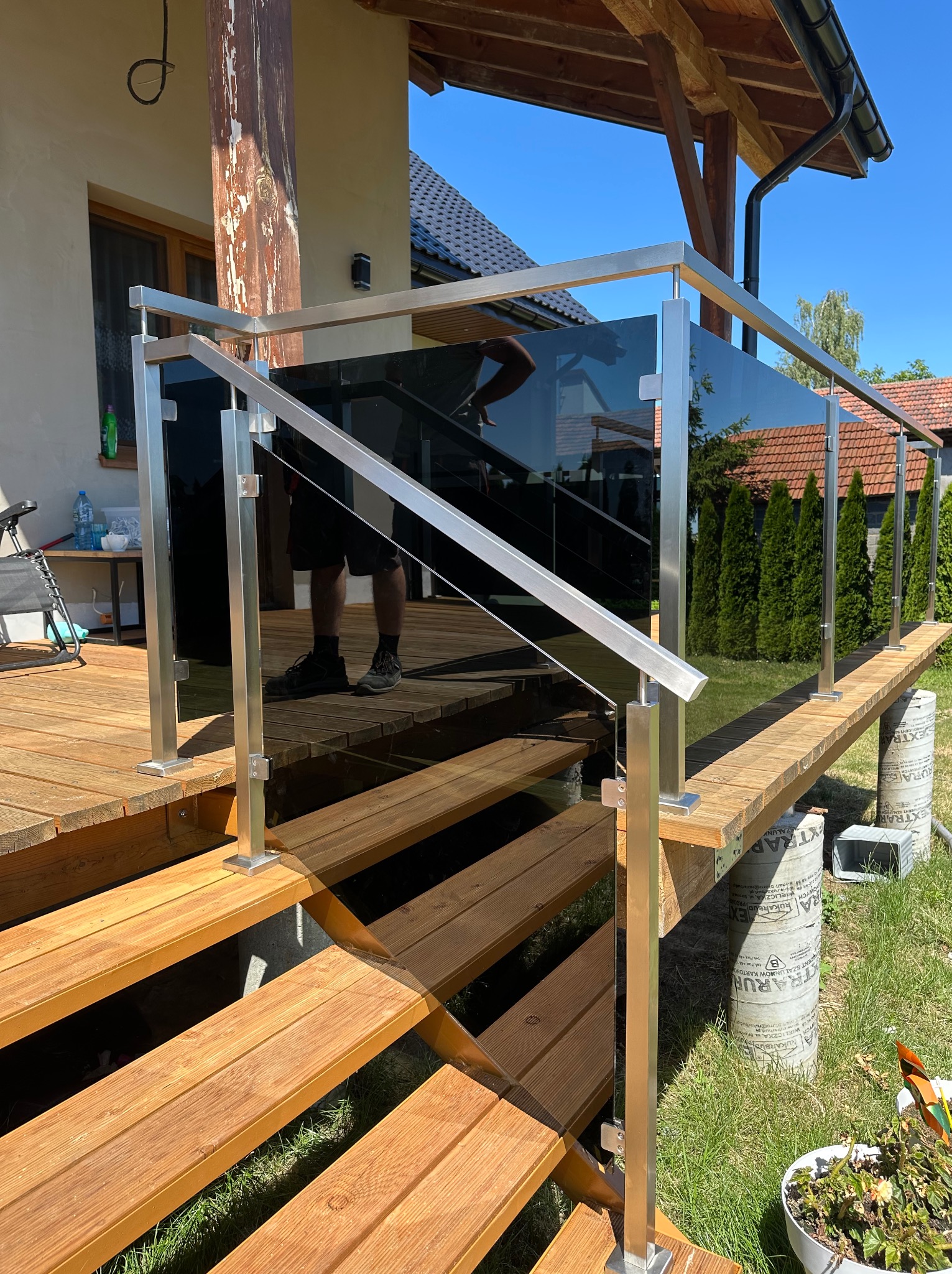 Szklana balustrada z metalowymi słupkami na drewnianym tarasie i schodach. Widoczne odbicie osoby. Tło: dom z ogrodem, słoneczny dzień.