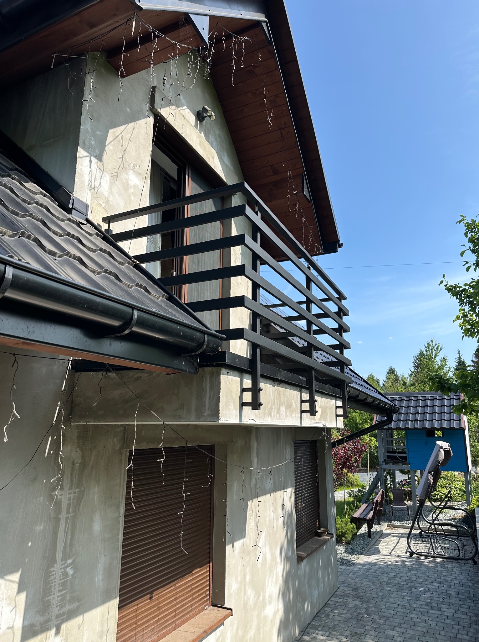 Czarna, pozioma balustrada balkonowa z metalu na tle jasnej elewacji domu i błękitnego nieba. Widok z dołu, oświetlenie słoneczne.