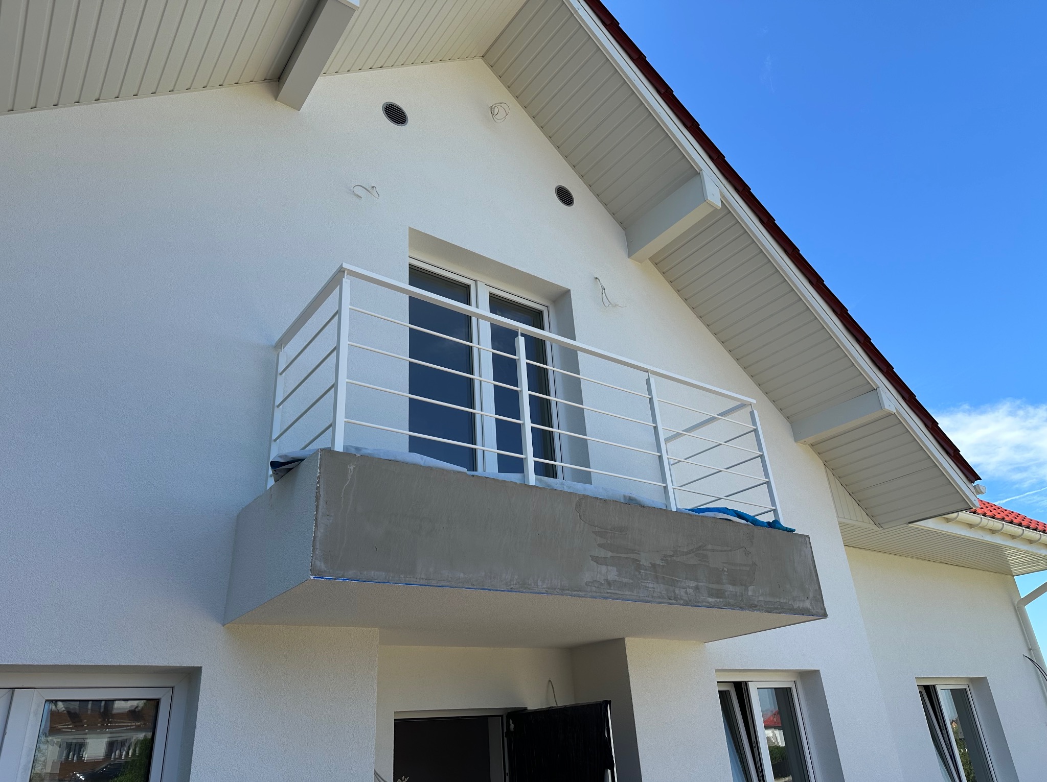 Biała balustrada balkonowa z poziomych prętów na tle białej elewacji domu z czerwonym dachem i błękitnym niebem. Detal architektoniczny.