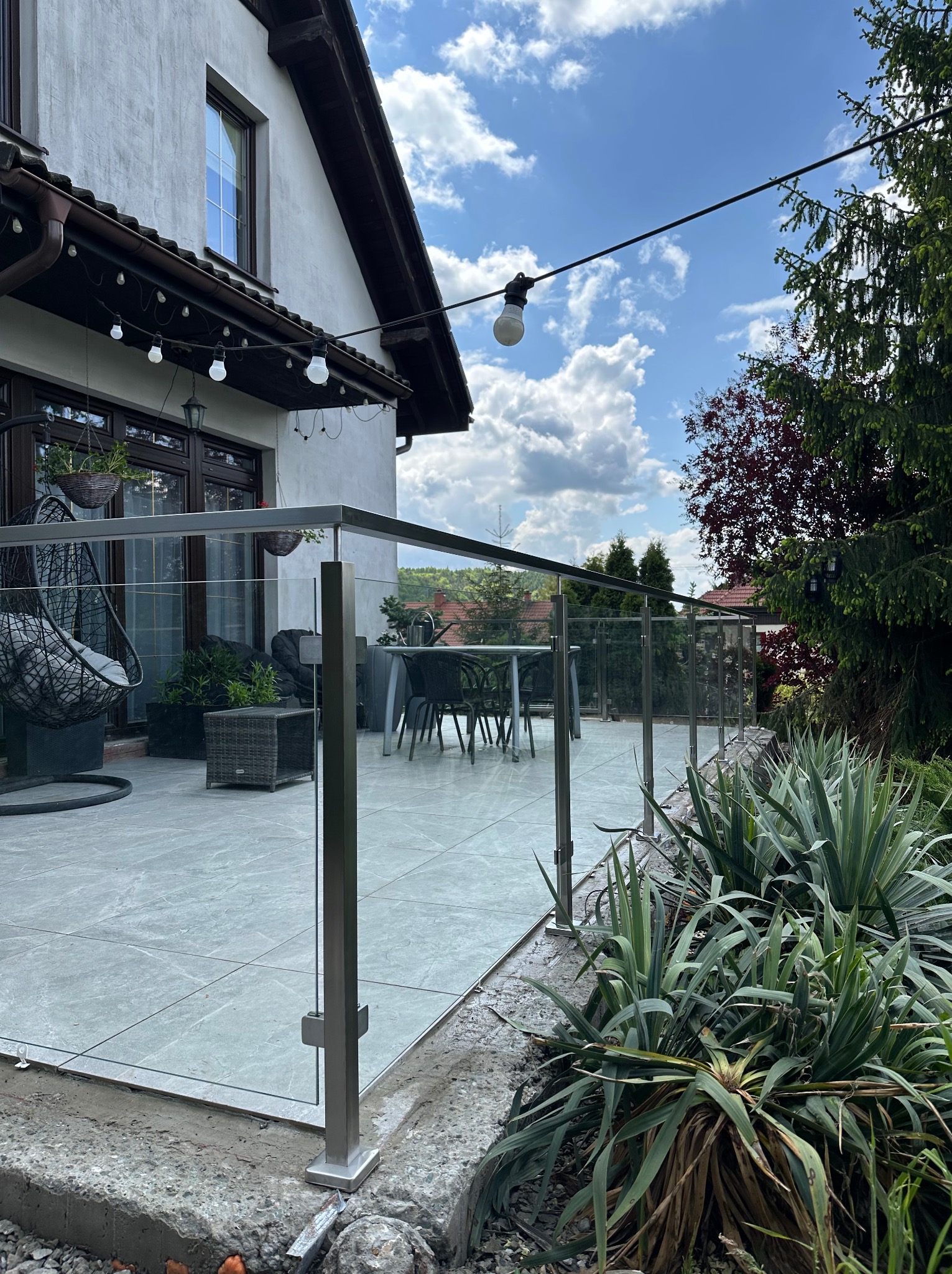 Szklana balustrada na tarasie z szarymi płytkami, widok na ogród i dom z dekoracyjnym oświetleniem. Nowoczesny design i eleganckie wykończenie.