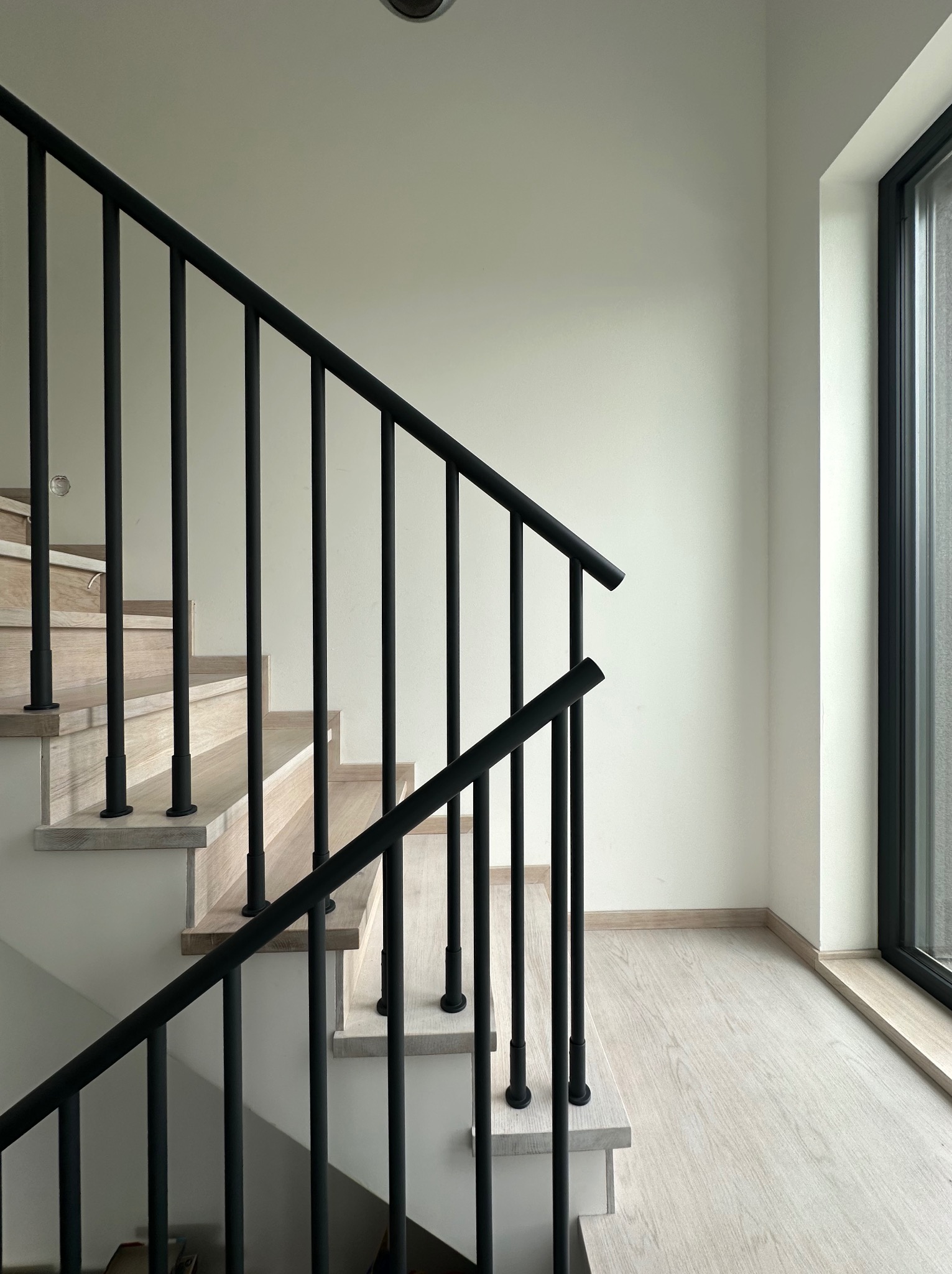Minimalistyczne schody z drewnianymi stopniami i czarną, metalową balustradą. Nowoczesny design, jasne wnętrze z oknem z czarną ramą. Prosta elegancja.