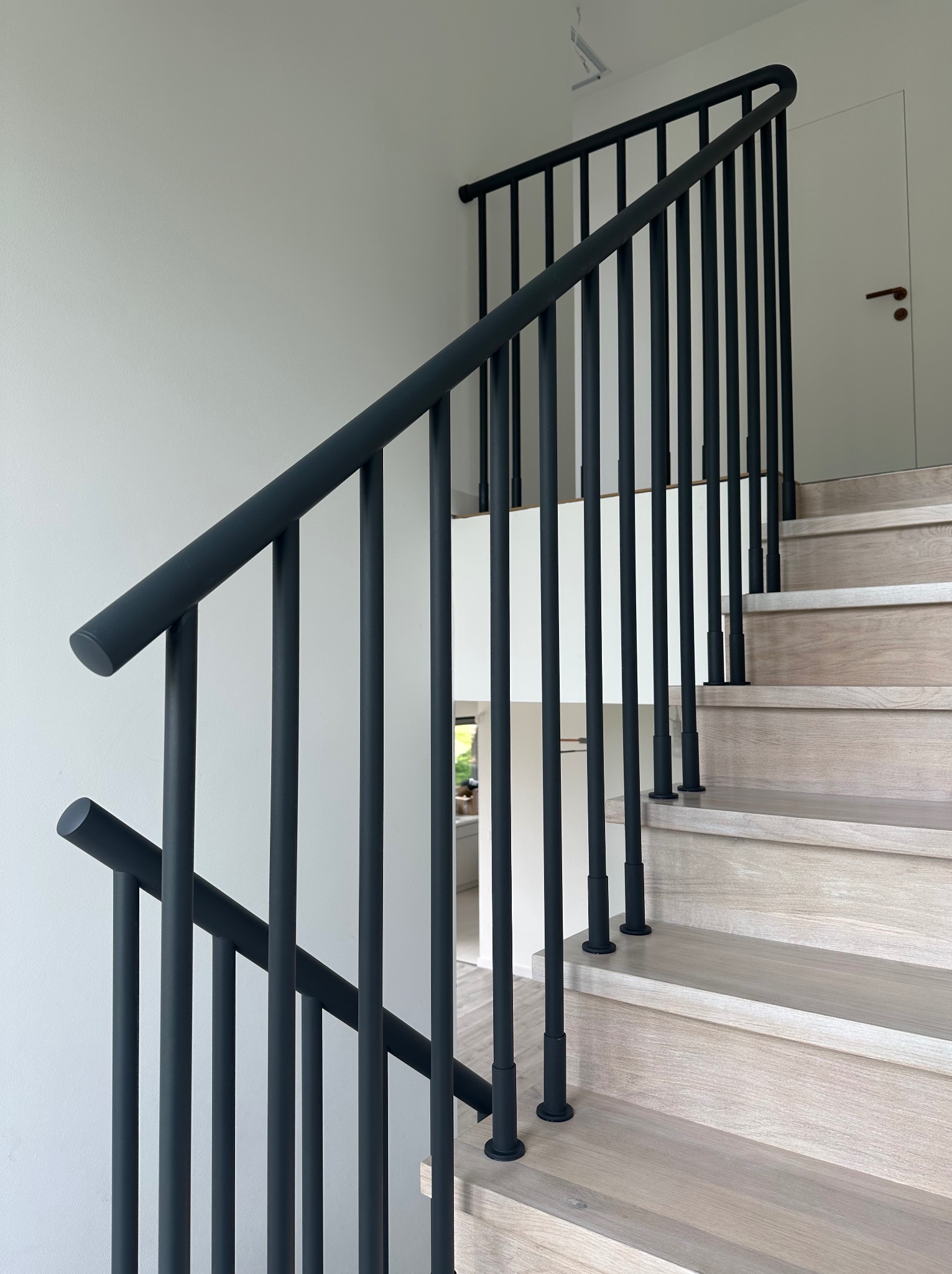 Nowoczesne schody z drewnianymi stopniami i minimalistyczną, ciemną balustradą z pionowych prętów. Widok z dołu, jasne tło.