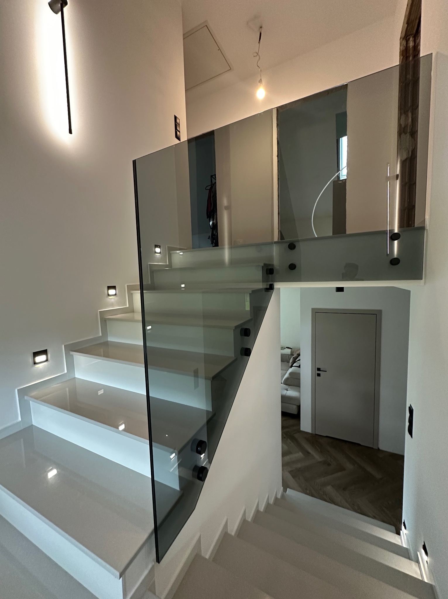 Nowoczesne schody wewnętrzne z białymi stopniami i szklaną balustradą, oświetlone punktowymi lampami ściennymi. W tle widoczny fragment salonu z parkietem.