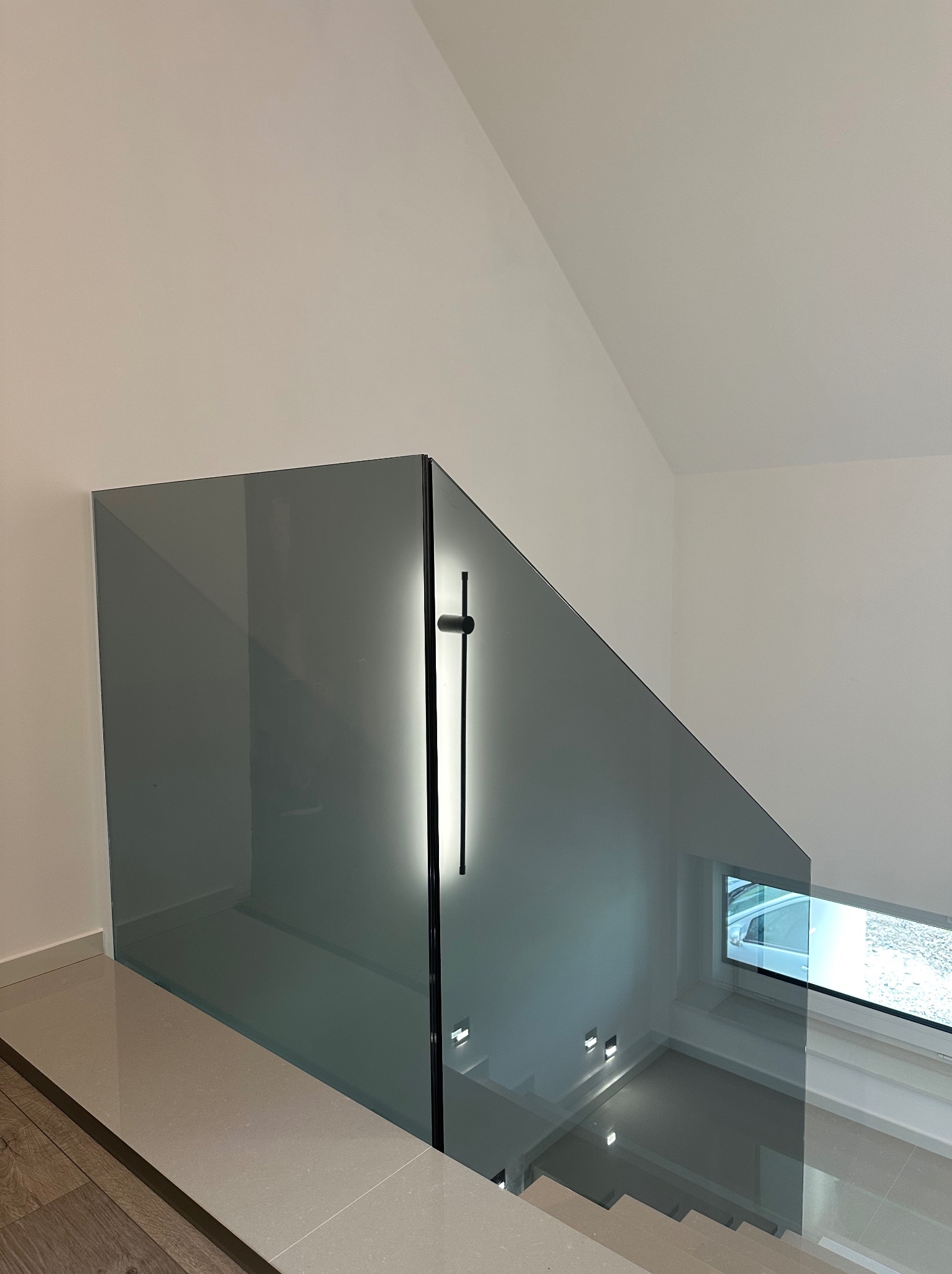 Szklana balustrada schodowa z oświetleniem LED w Wieliczce. Minimalistyczny design, widok z góry na schody z szarymi stopniami i okno z widokiem na zewnątrz.