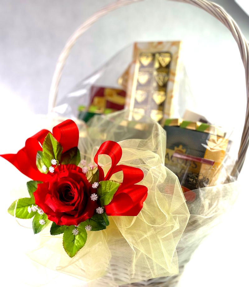 Wiklinowy kosz z czerwoną, materiałową różą, ozdobną wstążką i zawartością zapakowaną w przezroczystą folię.