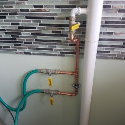 Gaz Instal Serwis - Usługi Gazownicze - Szymon Stefański - Miedziana instalacja z zaworami odcinającymi, biegnąca wzdłuż białej rury na tle ściany z mozaiką i zielonymi wężami.