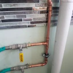 Miedziana instalacja wodna z zaworami KAS, zasilana elastycznymi przewodami w oplocie, zamontowana na tle ściany z mozaiką, widoczny fragment rury kanalizacyjnej.