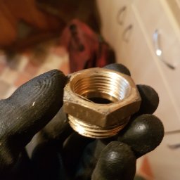 Gaz Instal Serwis - Usługi Gazownicze - Szymon Stefański - Mosiężna kształtka gwintowana trzymana w dłoni w czarnej rękawicy roboczej, widoczne ślady użytkowania na kształtce.