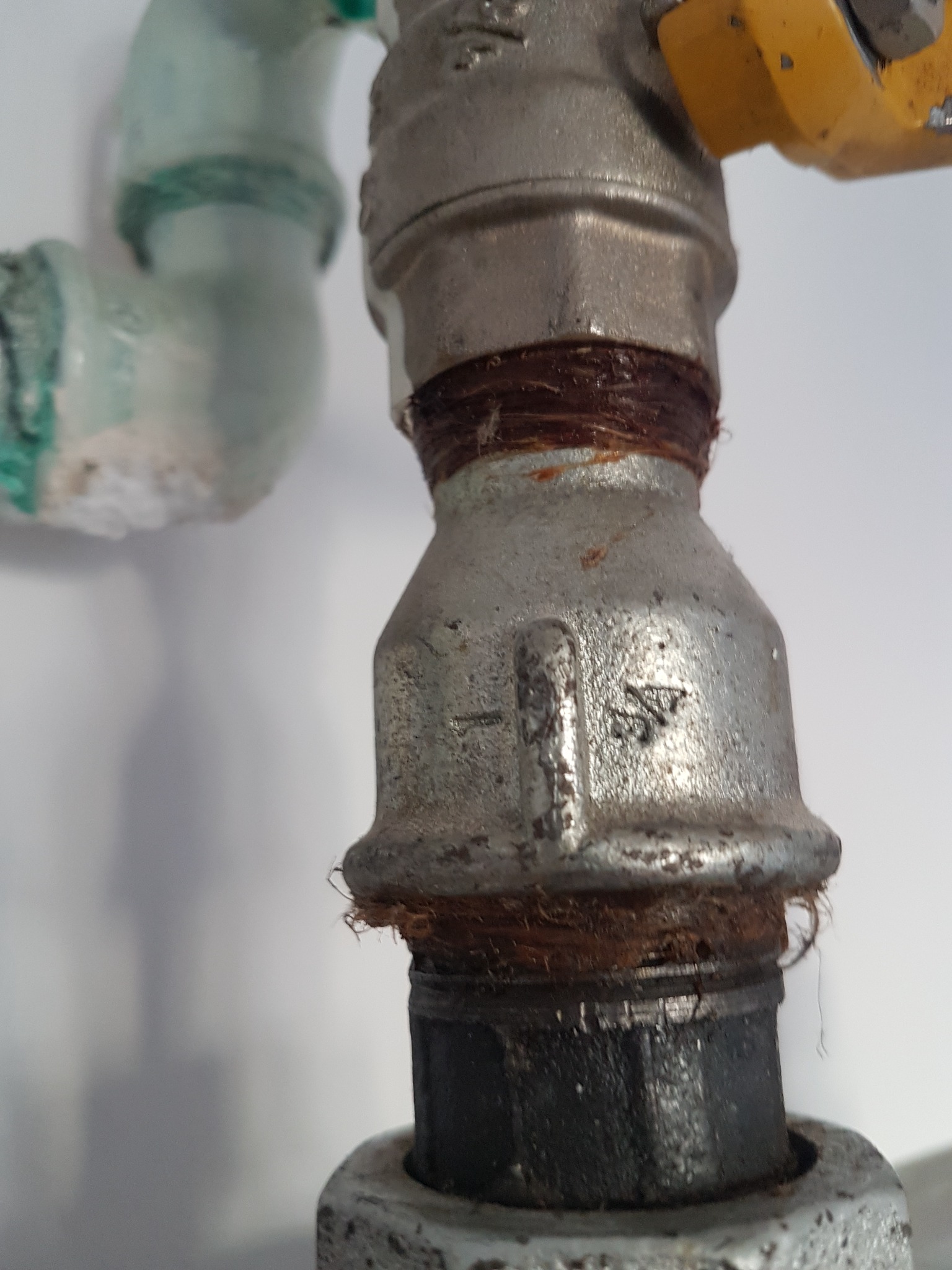 Metalowy zawór z widocznymi śladami uszczelniania konopiami, fragment instalacji gazowej lub wodnej z widocznymi oznaczeniami na korpusie.