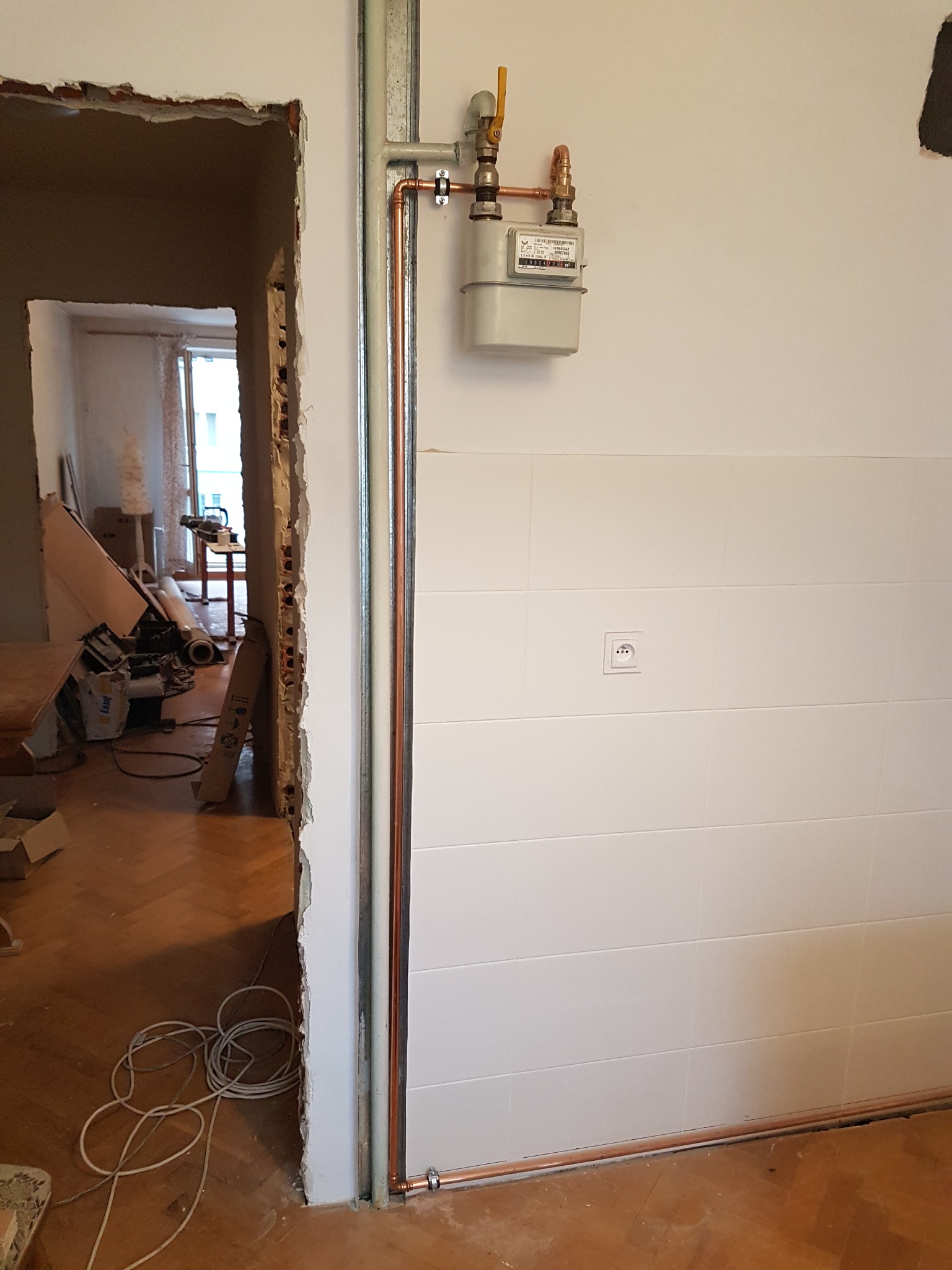 Nowa instalacja gazowa z licznikiem i miedzianymi rurami na białej ścianie z płytkami, widoczne gniazdko elektryczne i fragment remontowanego pomieszczenia z bałaganem w tle.