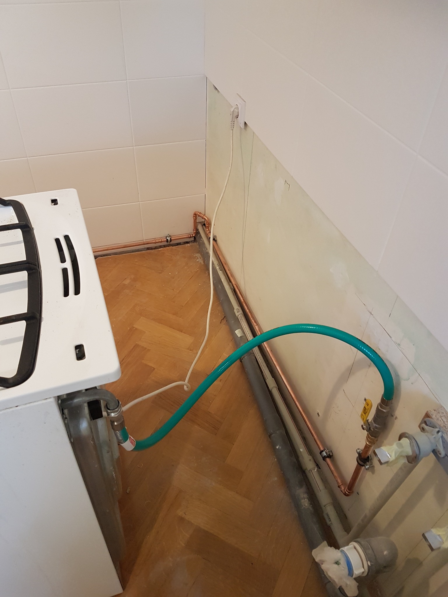 Podłączenie kuchenki gazowej elastycznym wężem do instalacji gazowej z zaworem odcinającym, widoczne rury miedziane i plastikowe, ściana częściowo wykończona płytkami.