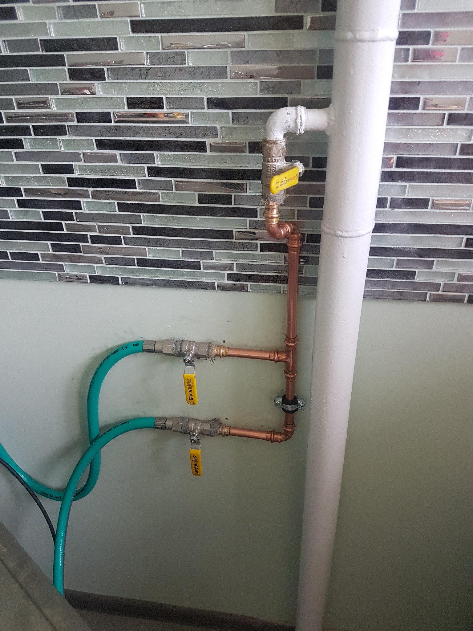 Miedziana instalacja z zaworami odcinającymi, biegnąca wzdłuż białej rury na tle ściany z mozaiką i zielonymi wężami.