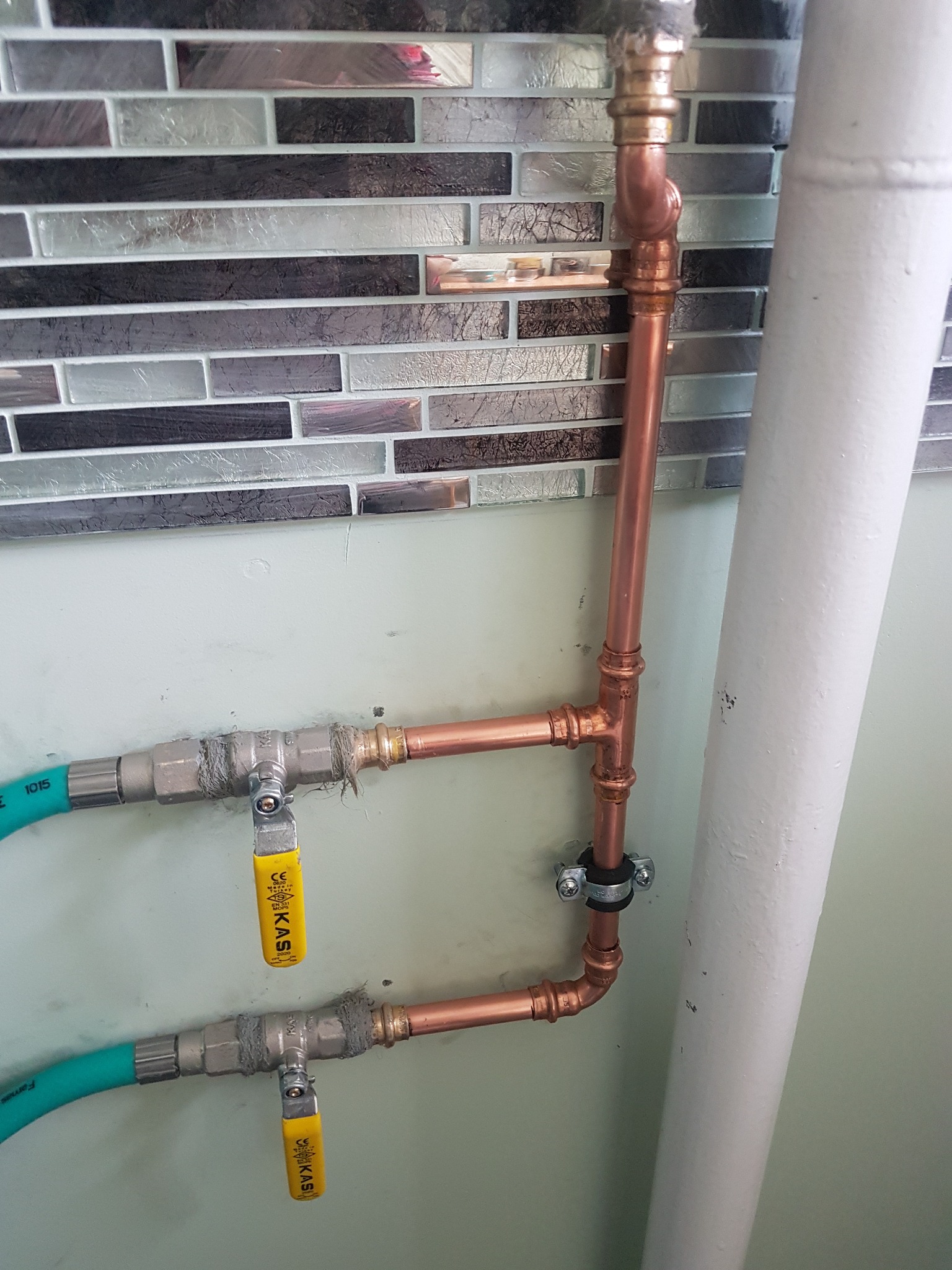 Miedziana instalacja wodna z zaworami KAS, zasilana elastycznymi przewodami w oplocie, zamontowana na tle ściany z mozaiką, widoczny fragment rury kanalizacyjnej.