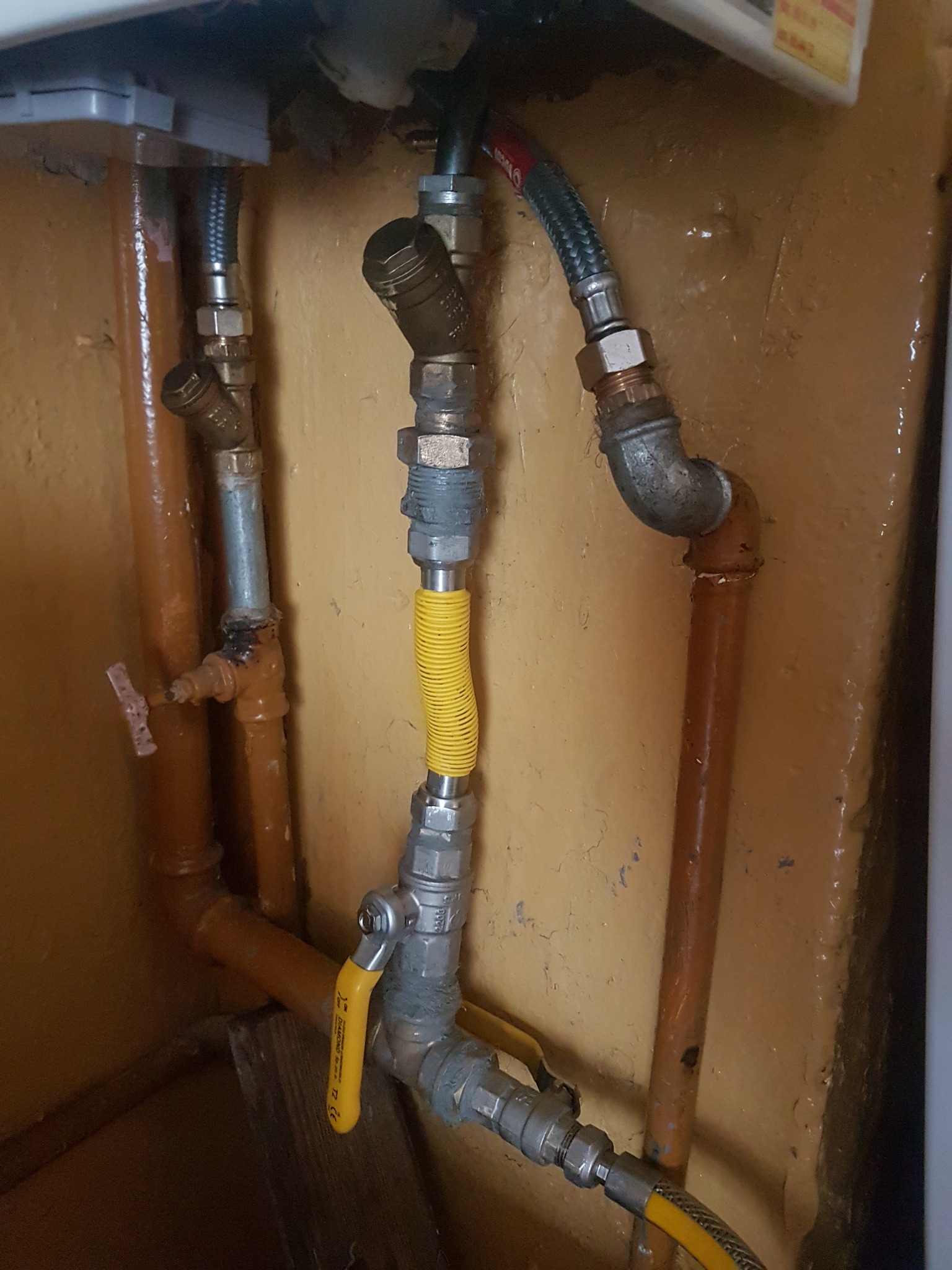 Fragment instalacji z rurami miedzianymi i elastycznym przewodem gazowym w żółtej osłonie, widoczne zawory odcinające i złączki, tło stanowią ściany w odcieniach żółci i brązu.