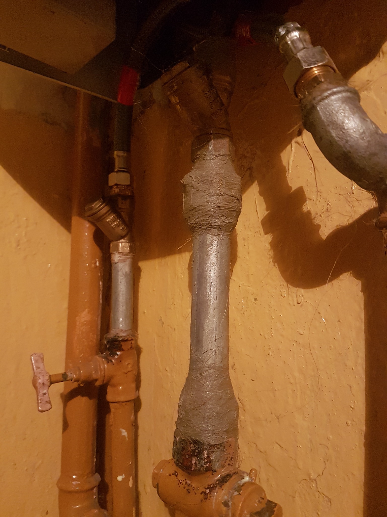Metalowe rury instalacji z zaworami, owinięte taśmą izolacyjną, na tle pomarańczowej ściany z widocznymi niedoskonałościami.