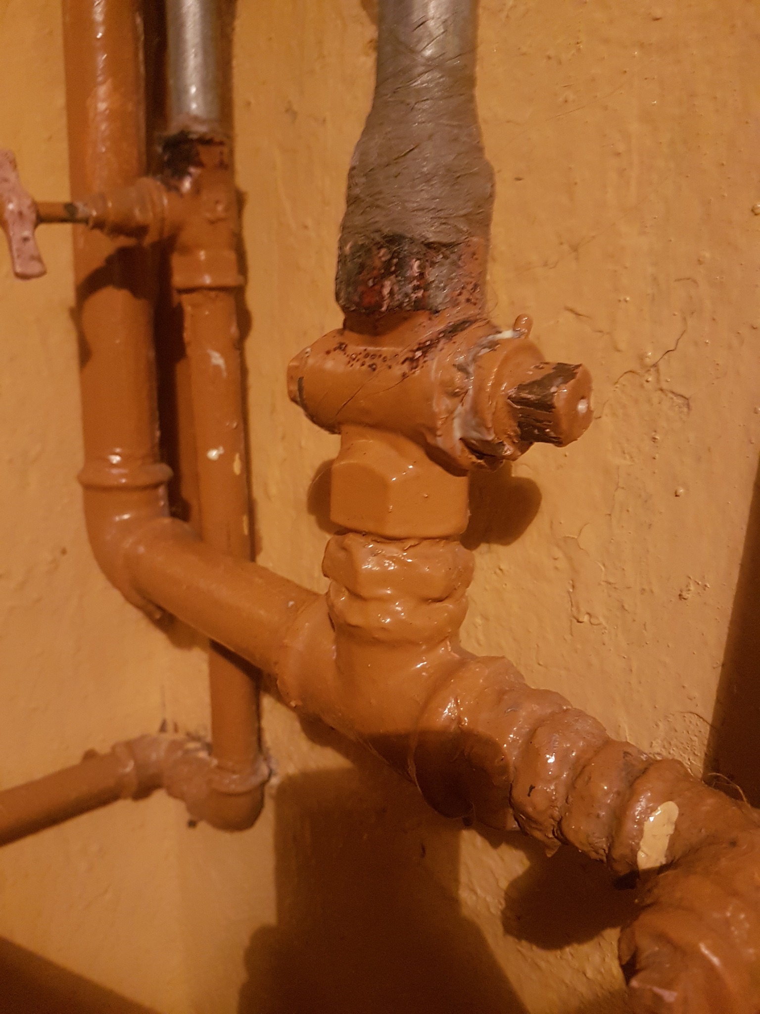 Brązowe rury gazowe z zaworem, pomalowane farbą, widoczne ślady użytkowania i niedokładnego malowania na tle pomarańczowej ściany.