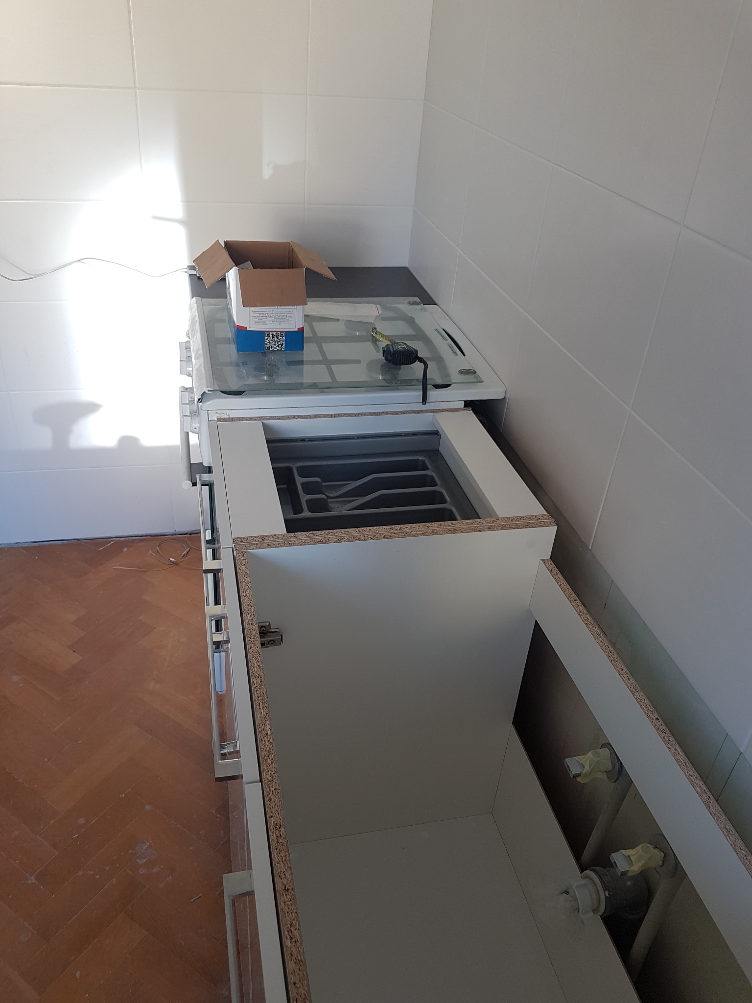 Wnętrze szafki pod zlew podczas montażu, widoczne rury instalacji wodnej, białe płytki na ścianie, podłoga z parkietu ułożonego w jodełkę oraz kartonowe pudełko na kuchence gazowej