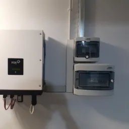 Biały inwerter marki FOX z podłączonymi kablami, zamontowany na ścianie obok dwóch szarych skrzynek rozdzielczych marki Noark z przezroczystymi pokrywami.