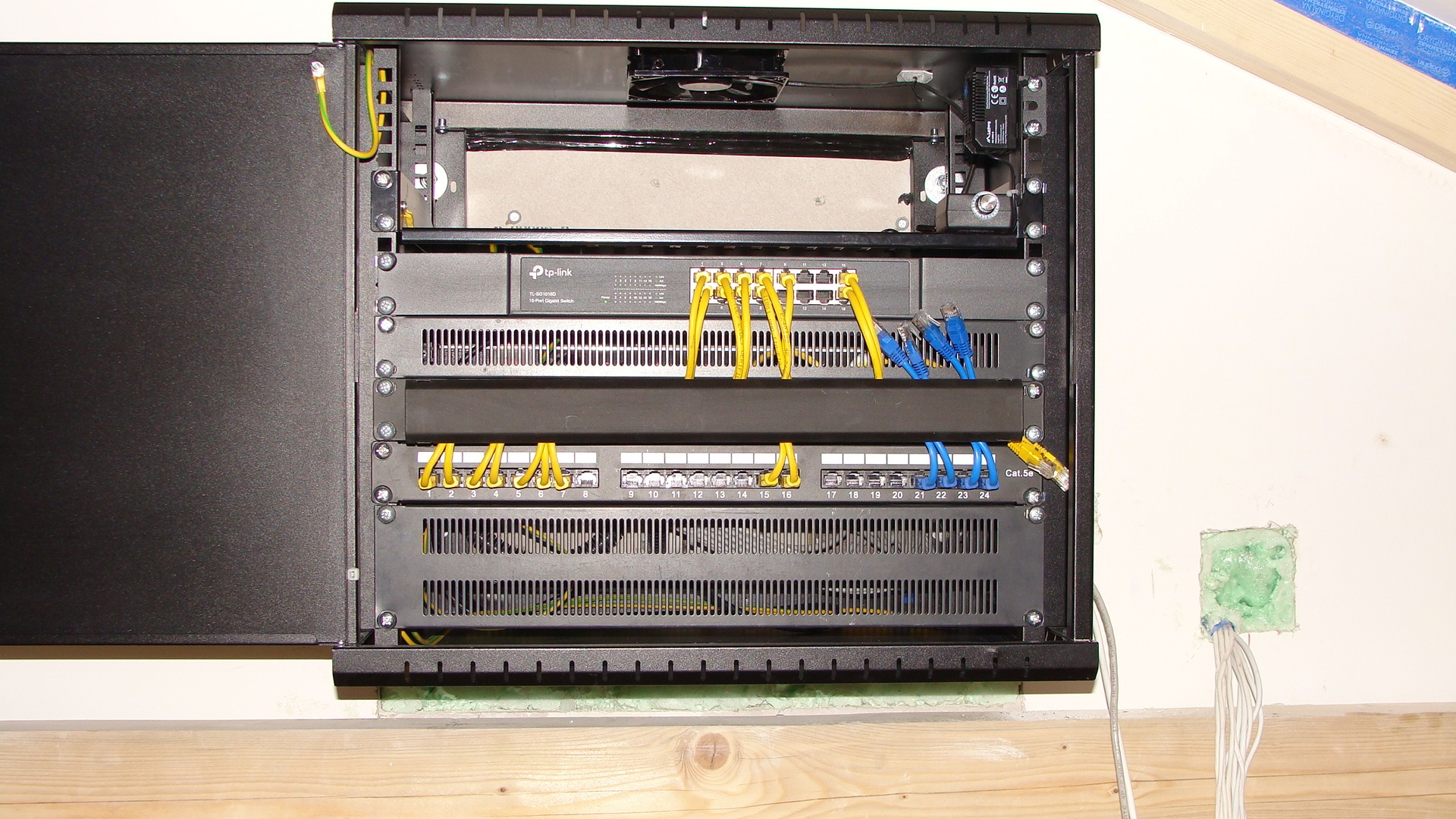 Zamknięta szafa rack 19 cali z zamontowanym switchem TP-Link, patch panelem i okablowaniem UTP w kolorach żółtym i niebieskim, zasilana PoE, zamontowana na ścianie z widoczną instalacją elektryczną...