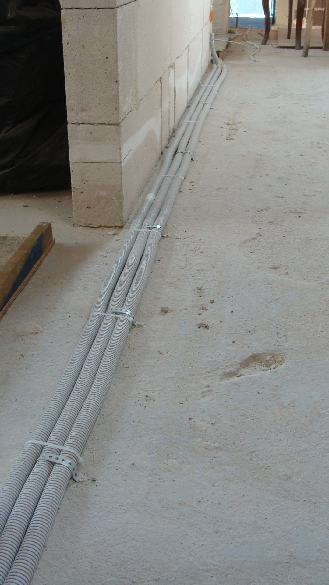 Trzy szare rury karbowane prowadzone wzdłuż ściany z betonu komórkowego na surowym betonowym podłożu, mocowane metalowymi uchwytami i białymi opaskami zaciskowymi.