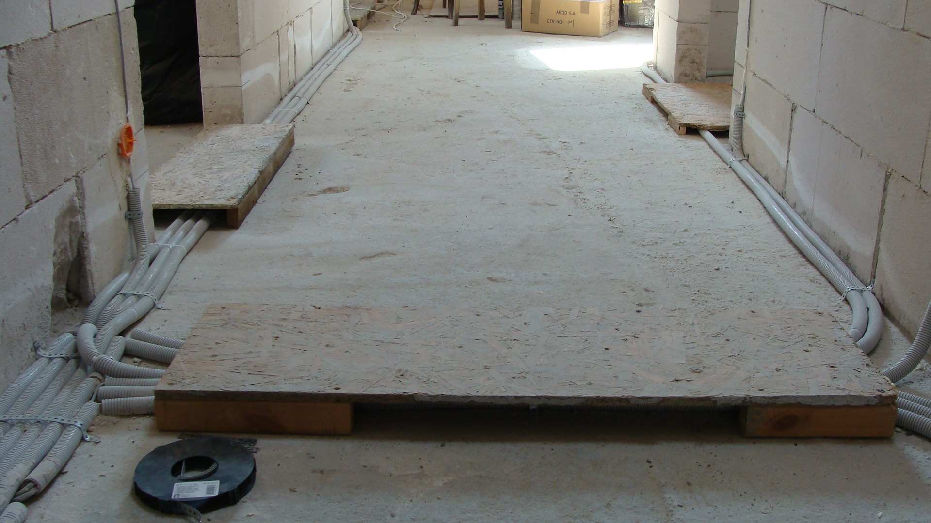 Szare rury instalacji elektrycznej prowadzone wzdłuż ścian surowego pomieszczenia, częściowo ukryte pod drewnianymi podestami, na podłodze leży zwój taśmy instalacyjnej.