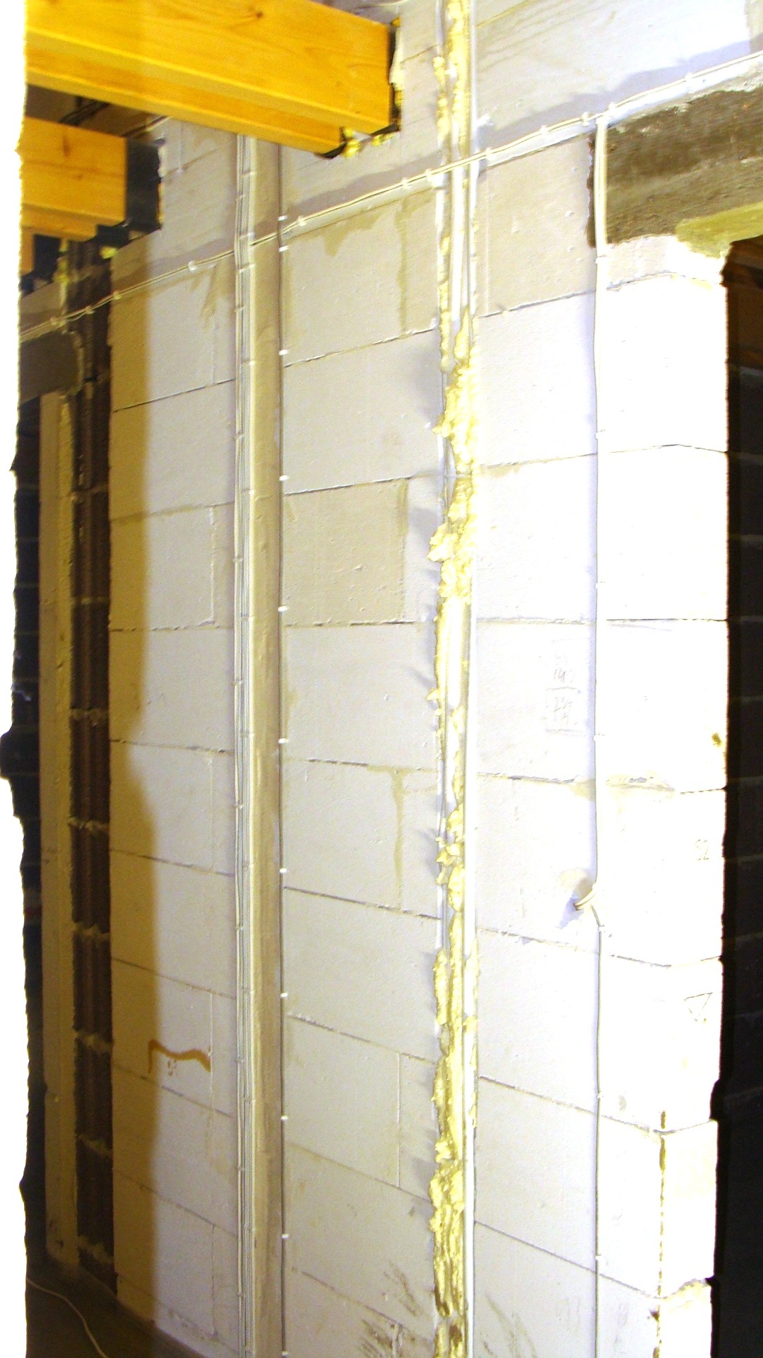 Ściana z betonu komórkowego w trakcie prac wykończeniowych, widoczne kable elektryczne prowadzone wzdłuż ściany i zabezpieczone pianką montażową, drewniane belki stropowe u góry kadru.