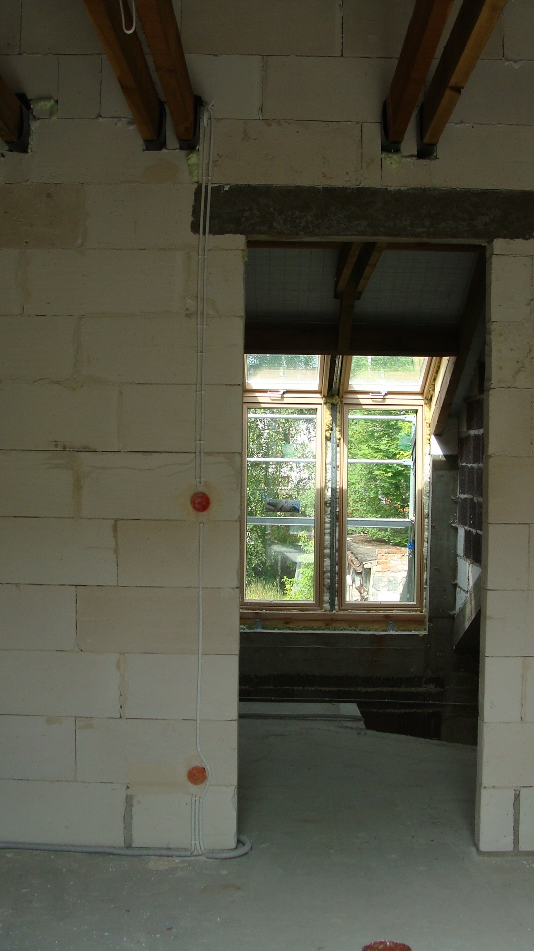 Surowy stan budynku w trakcie prac wykończeniowych: widoczne okno dachowe, ściany z betonu komórkowego z puszkami instalacyjnymi i przewodami elektrycznymi, drewniane belki stropowe.