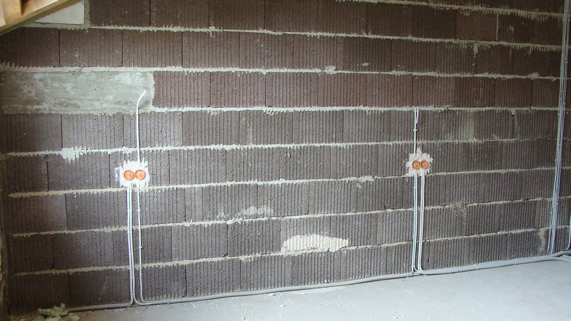 Surowa ściana z szarego betonu komórkowego z widocznymi przewodami elektrycznymi i puszkami podwójnych gniazdek, przygotowana do tynkowania.