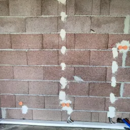 Surowa ściana z betonu komórkowego z wywierconymi otworami i pomarańczowymi puszkami instalacyjnymi, częściowo zamaskowanymi gipsem, widoczne przewody elektryczne i rury instalacyjne na podłodze.