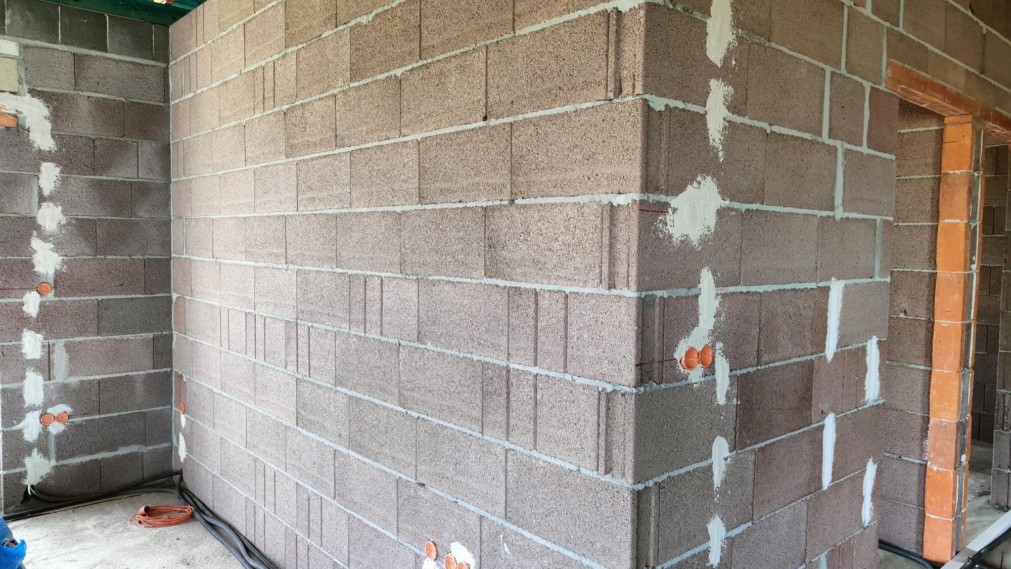 Surowe ściany z bloczków betonu komórkowego w trakcie budowy, widoczne pomarańczowe puszki instalacyjne w ścianie, szpachlowane miejsca wokół puszek i spoin, przewody elektryczne na podłodze.