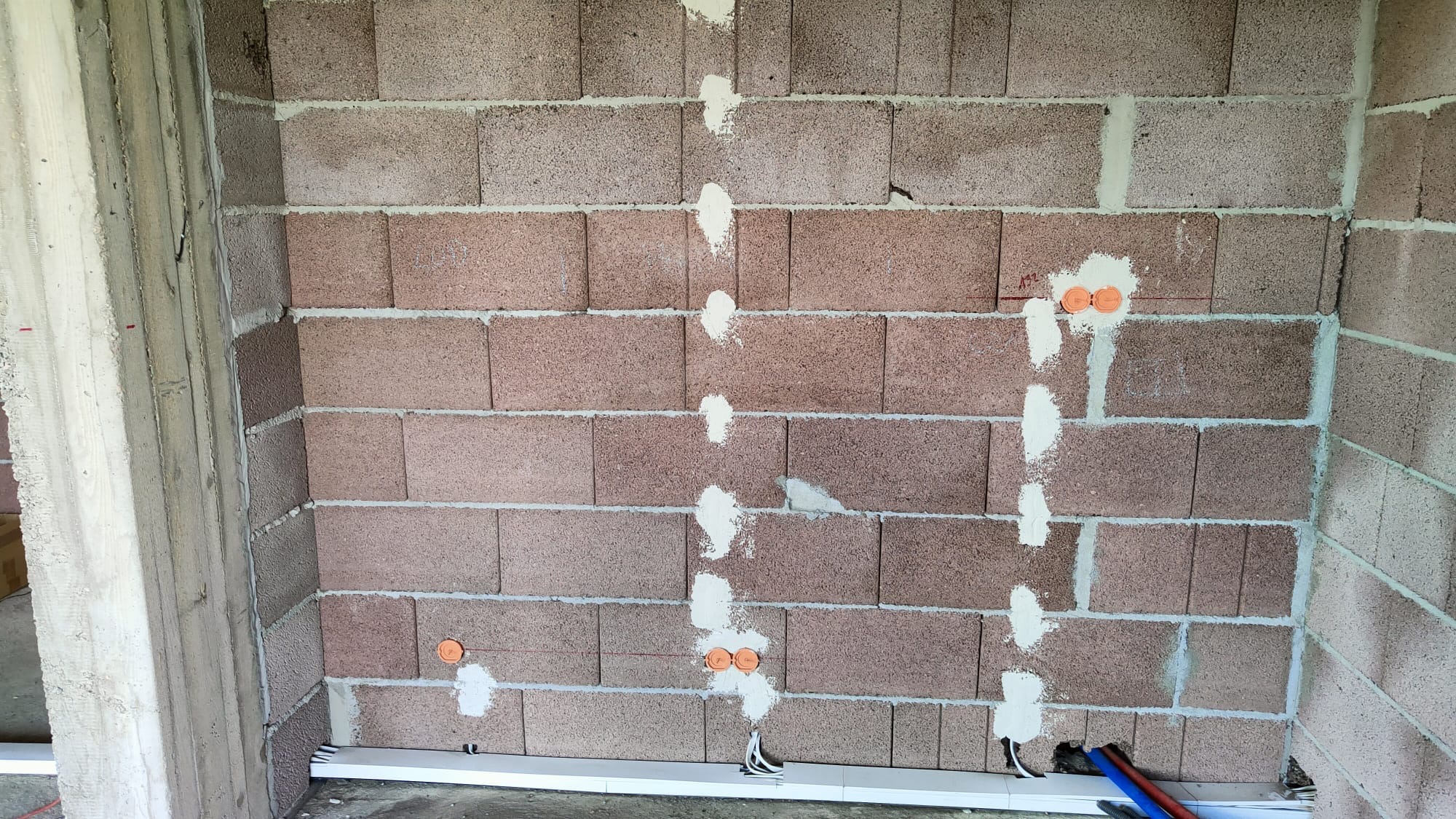 Surowa ściana z betonu komórkowego z wywierconymi otworami i pomarańczowymi puszkami instalacyjnymi, częściowo zamaskowanymi gipsem, widoczne przewody elektryczne i rury instalacyjne na podłodze.