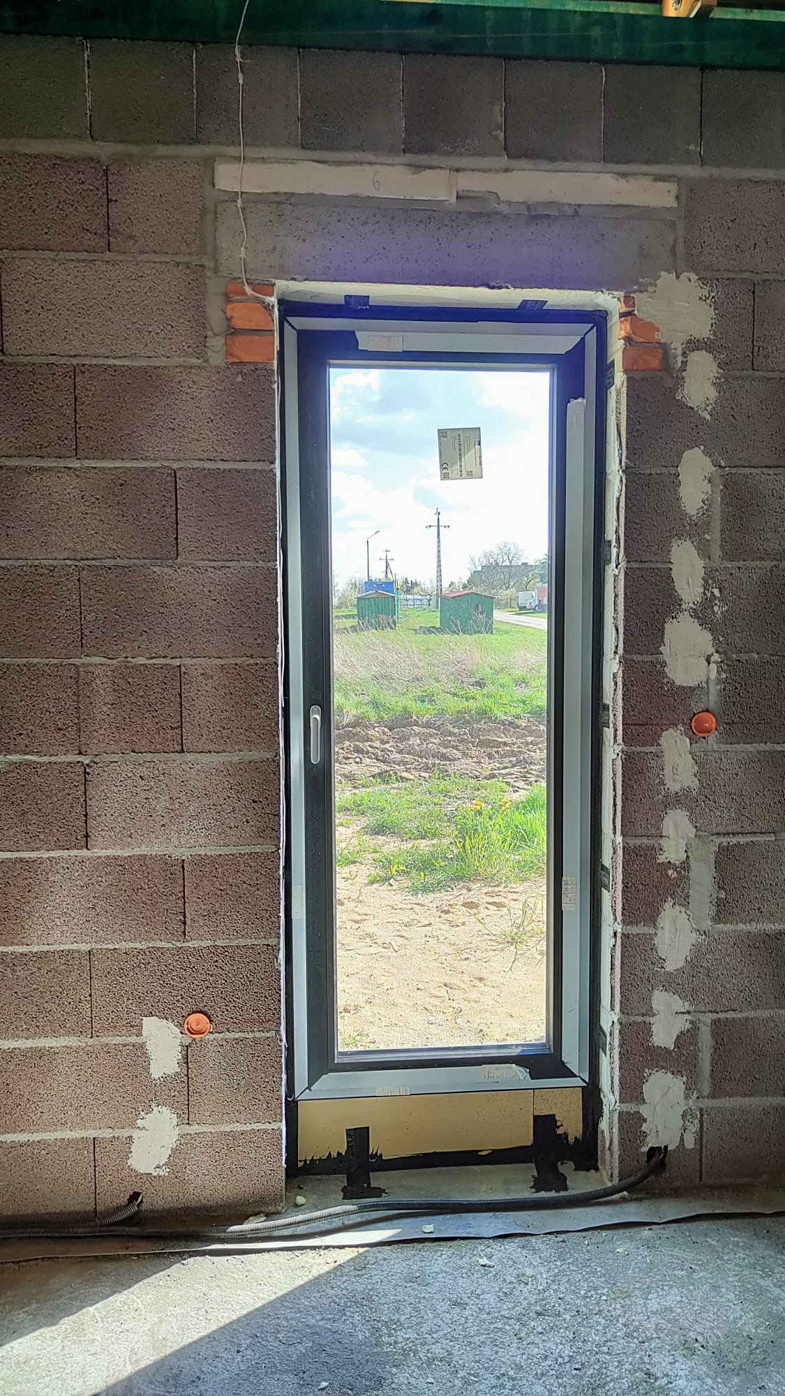 Wstawione okno PCV w surowym murze z betonu komórkowego, widoczne pomarańczowe puszki instalacyjne i przewody elektryczne w peszlach, widok na podwórko z zielonymi budami przez szybę.