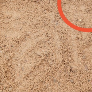 Beżowa pospółka piaskowa o zróżnicowanej granulacji, widoczne drobne kamyczki i grudki ziemi, oświetlona naturalnym światłem z góry, z widocznym elementem graficznym w prawym górnym rogu.