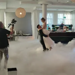 Uśmiechnięty pan młody w czarnym garniturze trzyma na rękach pannę młodą w białej sukni, otoczeni sztuczną mgłą podczas pierwszego tańca, filmowani przez operatora z kamerą na stabilizatorze