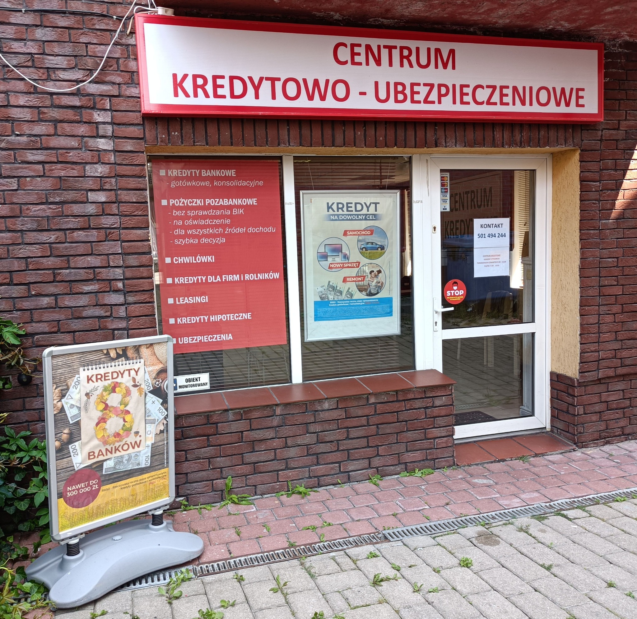 Fasada budynku Centrum Kredytowo-Ubezpieczeniowego z szyldem, plakatem w oknie i stojakiem reklamowym na chodniku.